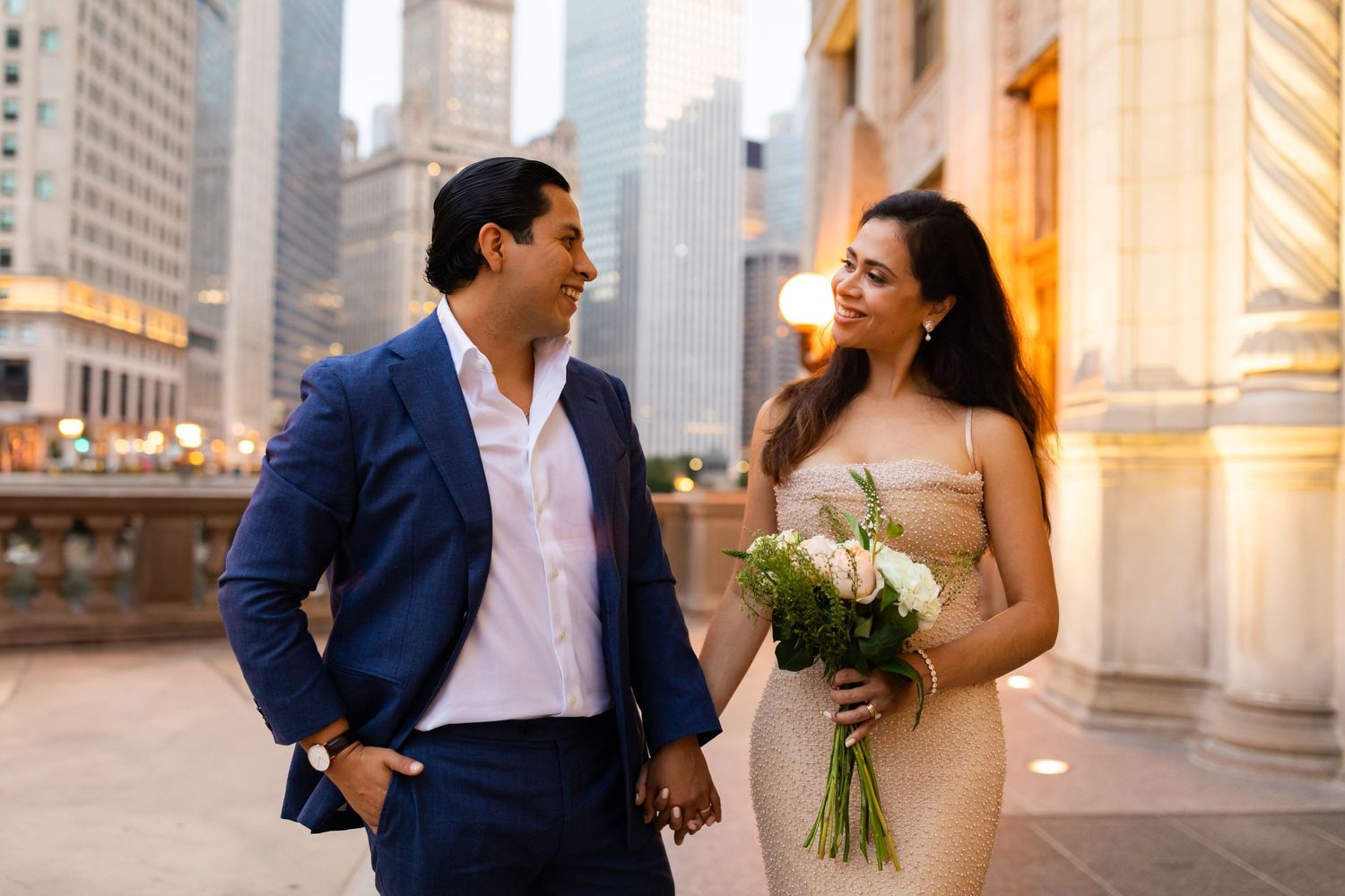 Bride in gold dress and groom in navy suit hold hands smiling in Chicago cityscape at dusk.