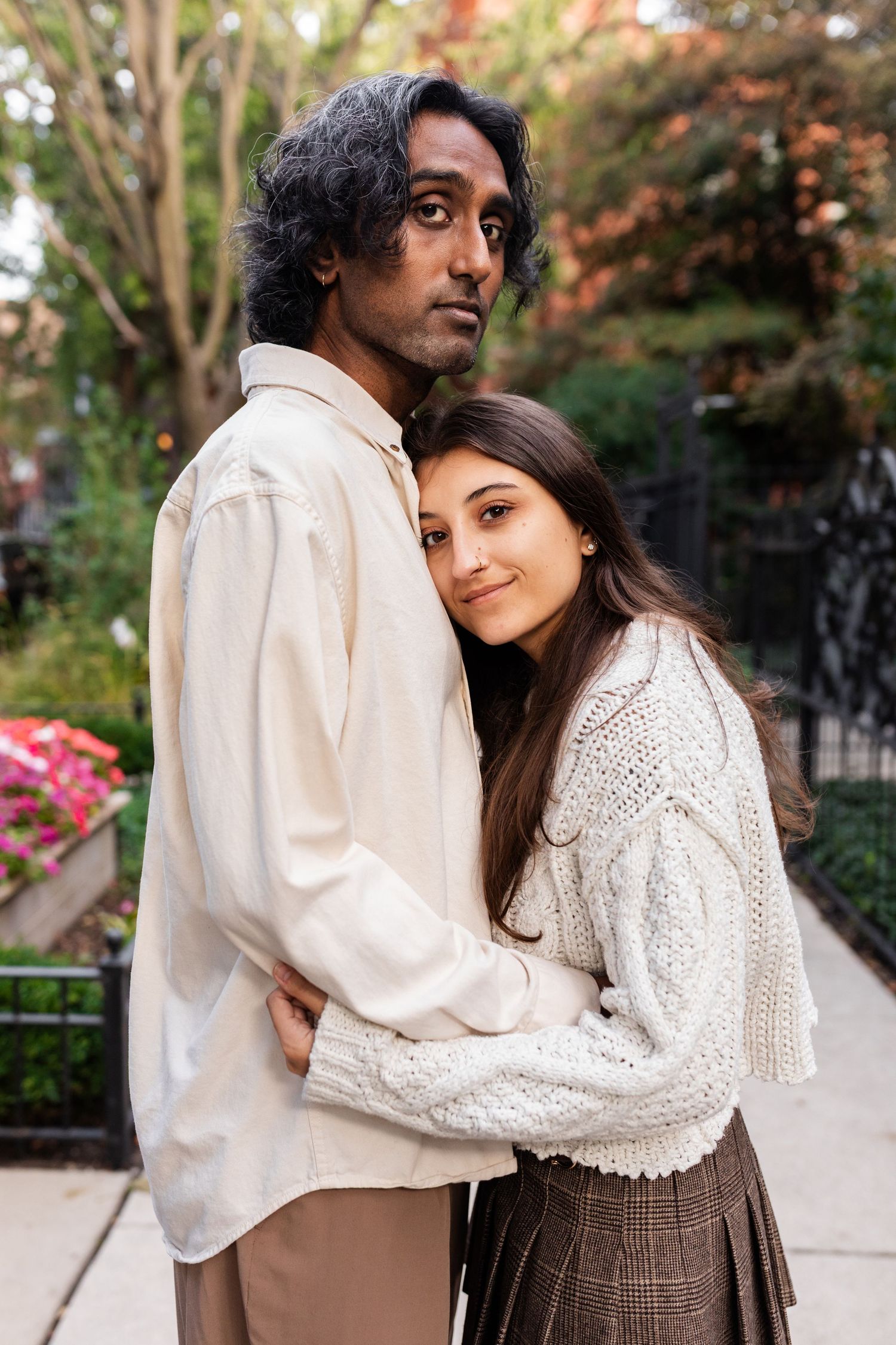 Interracial couple embracing on an urban sidewalk, woman in cream knit sweater and plaid skirt leaning against man in white shirt.
