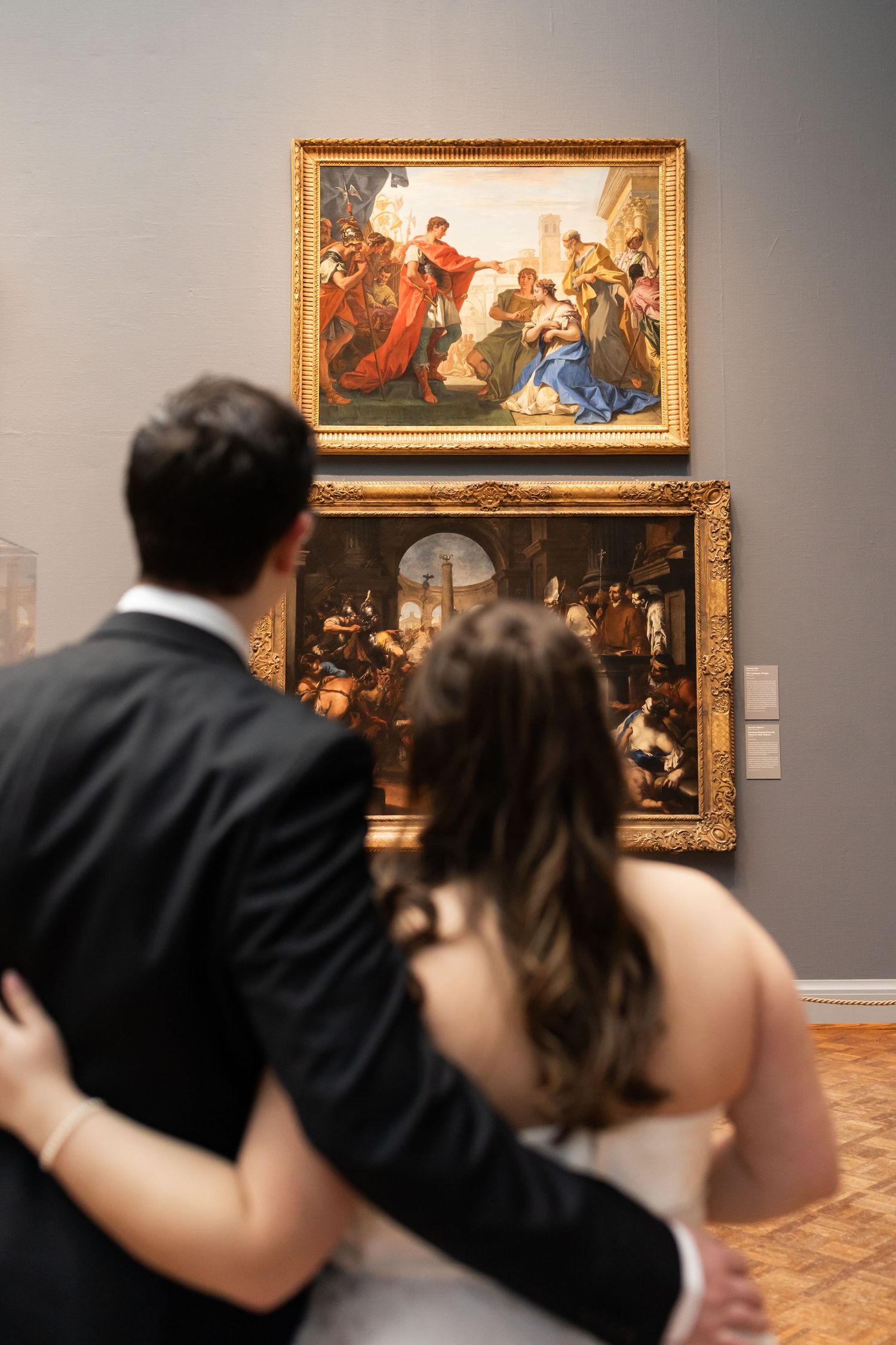 Newlyweds admire classical paintings together in ornate gold frames at art museum gallery.