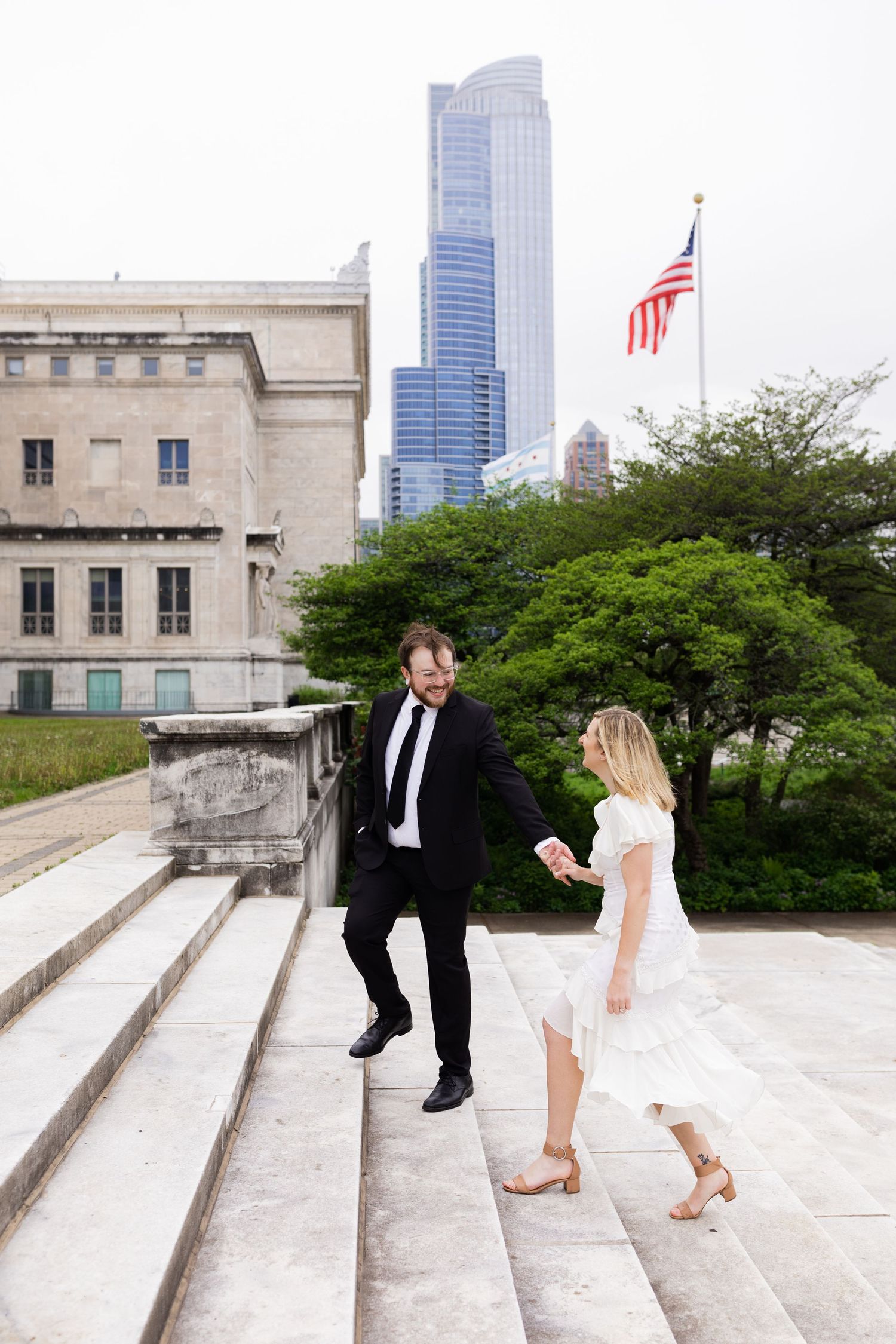 Groom leads bride up grand marble steps with American flag and city skyscrapers in the background.