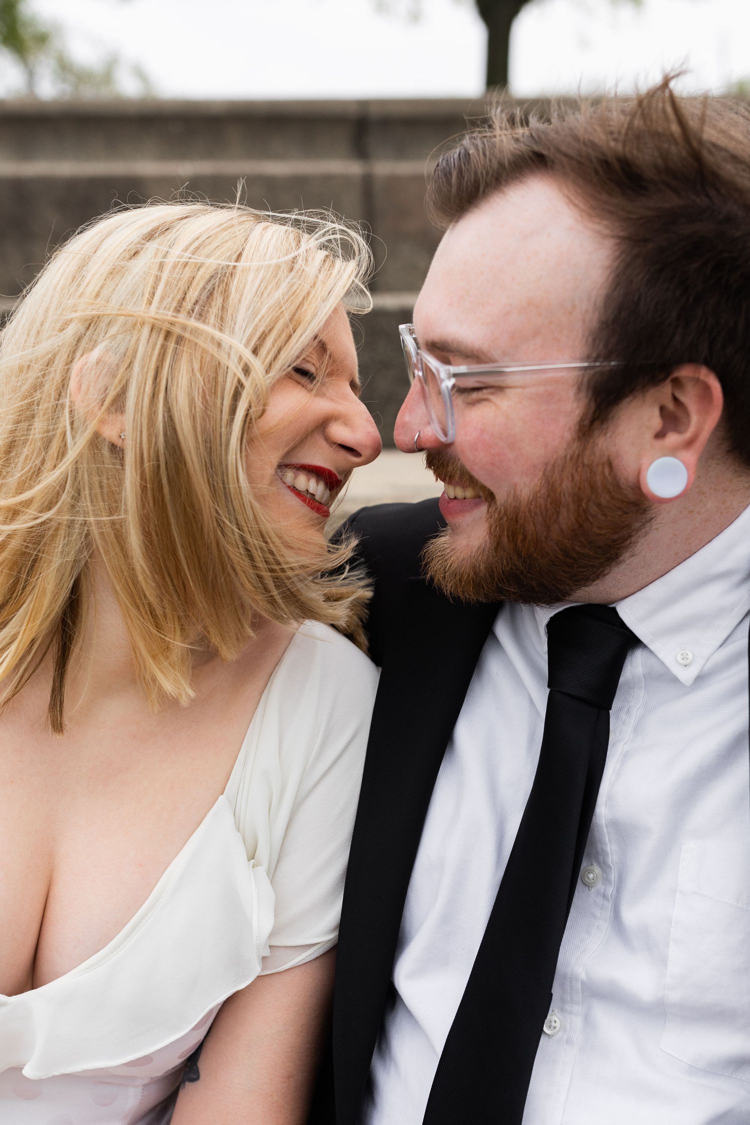 A laughing couple looks at each other closely outdoors, the woman with blonde hair and the man with a beard.
