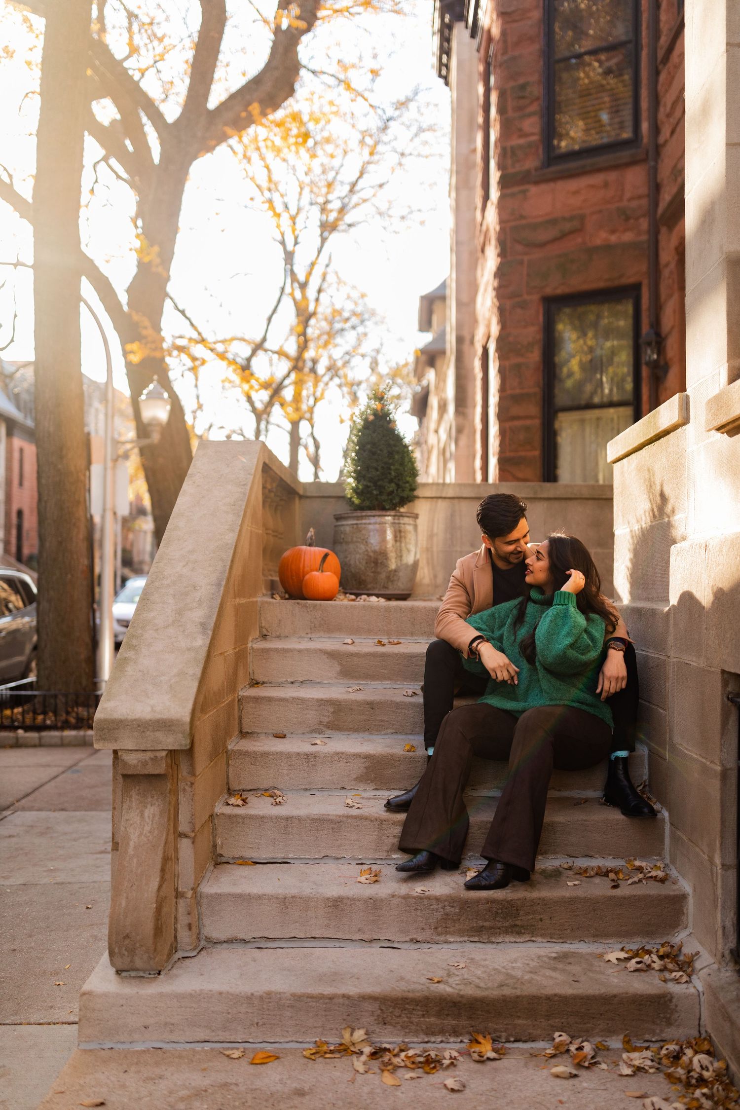 A couple sitting together on urban brownstone steps surrounded by autumn leaves and a Halloween pumpkin.