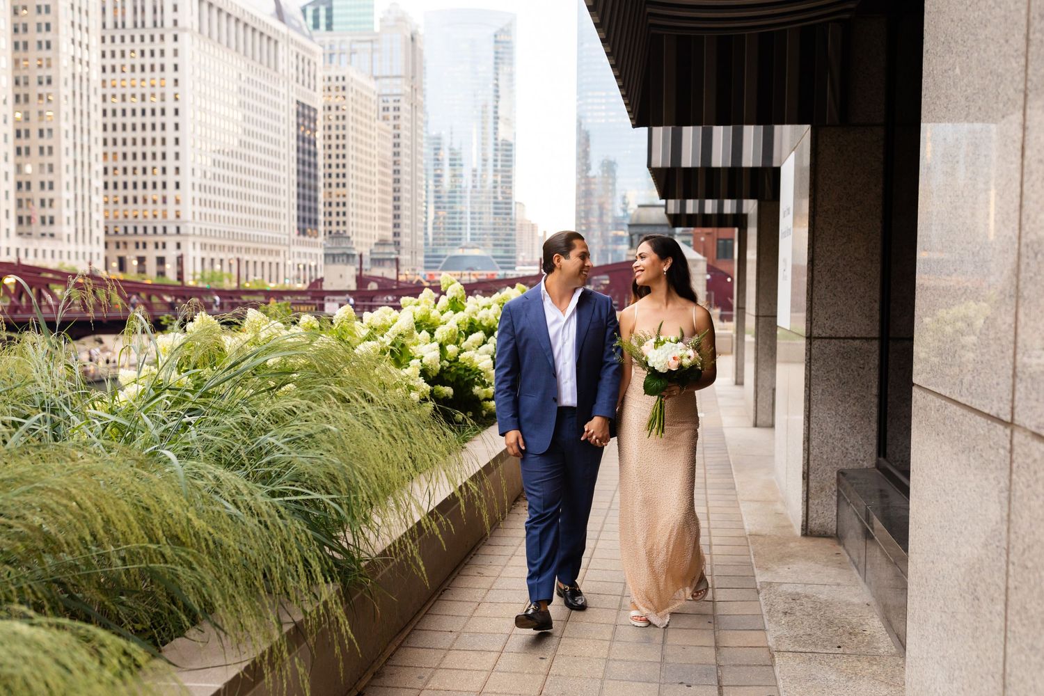 Bride and groom walk hand in hand along Chicago Riverwalk with city skyline in background.