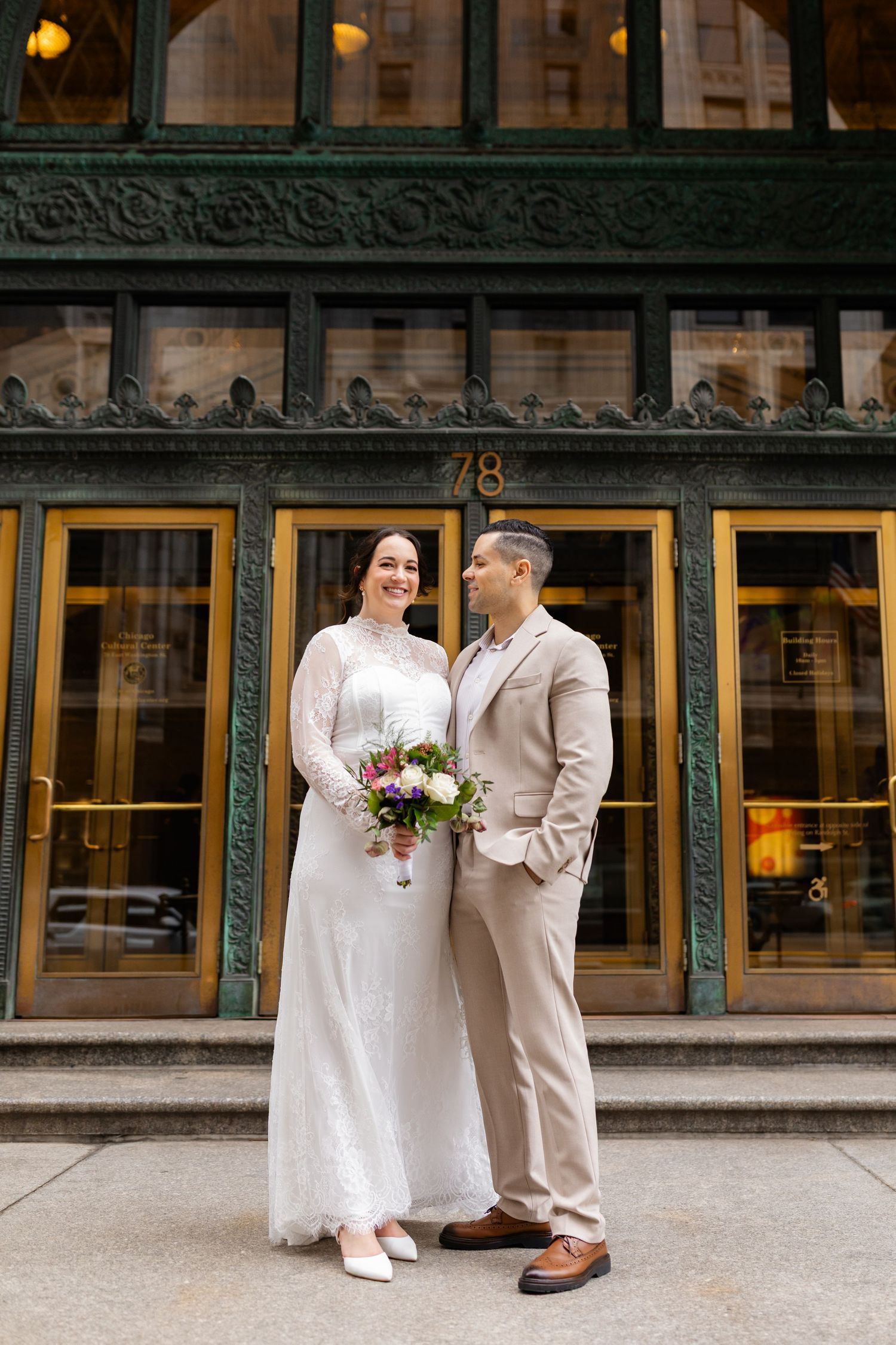 Elegant bride and groom standing together outside ornate building 78, bride holding wildflower bouquet.