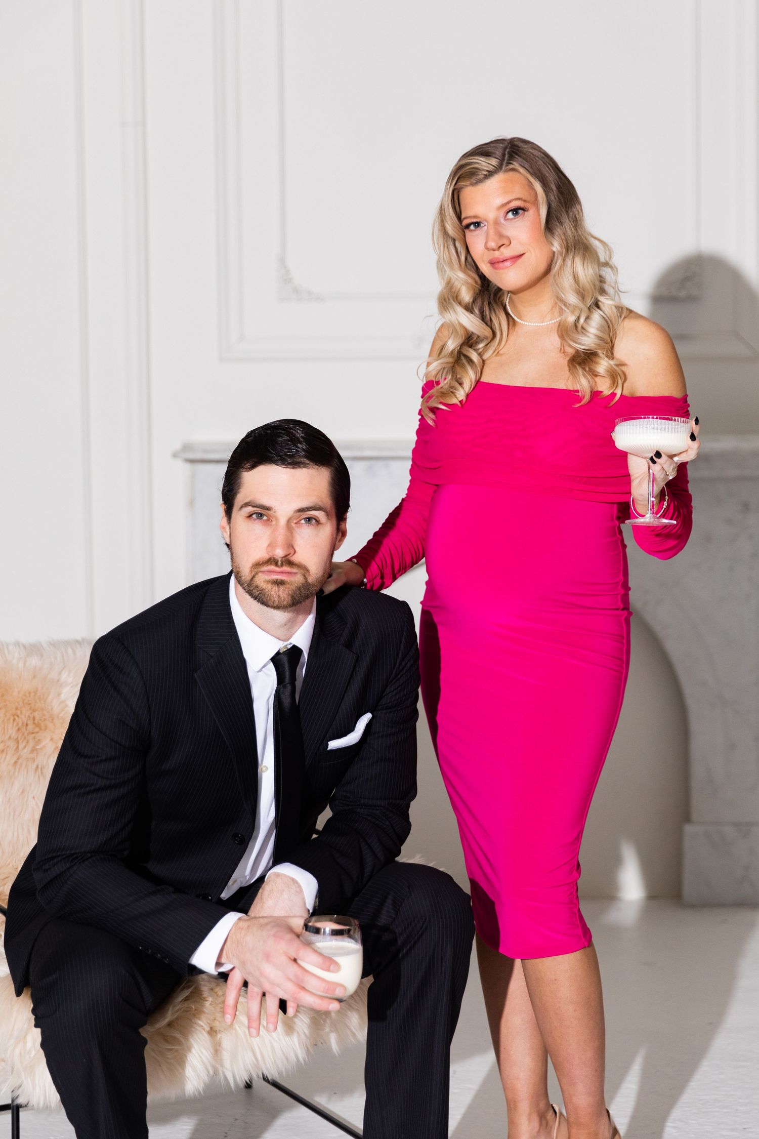 Stylish couple in a white studio, man in black suit seated and woman in pink off-shoulder dress standing.