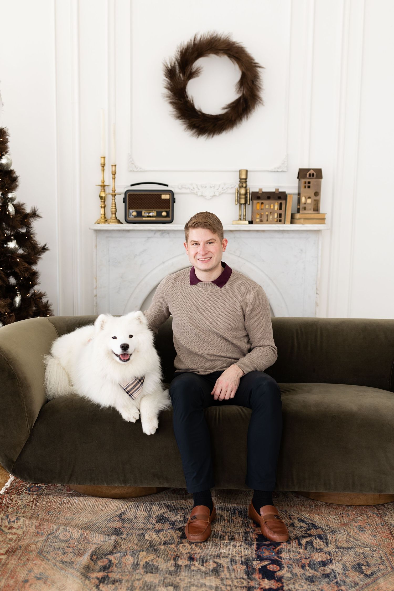 Smiling young man sitting on green velvet sofa with white Samoyed dog in Christmas decorated room.