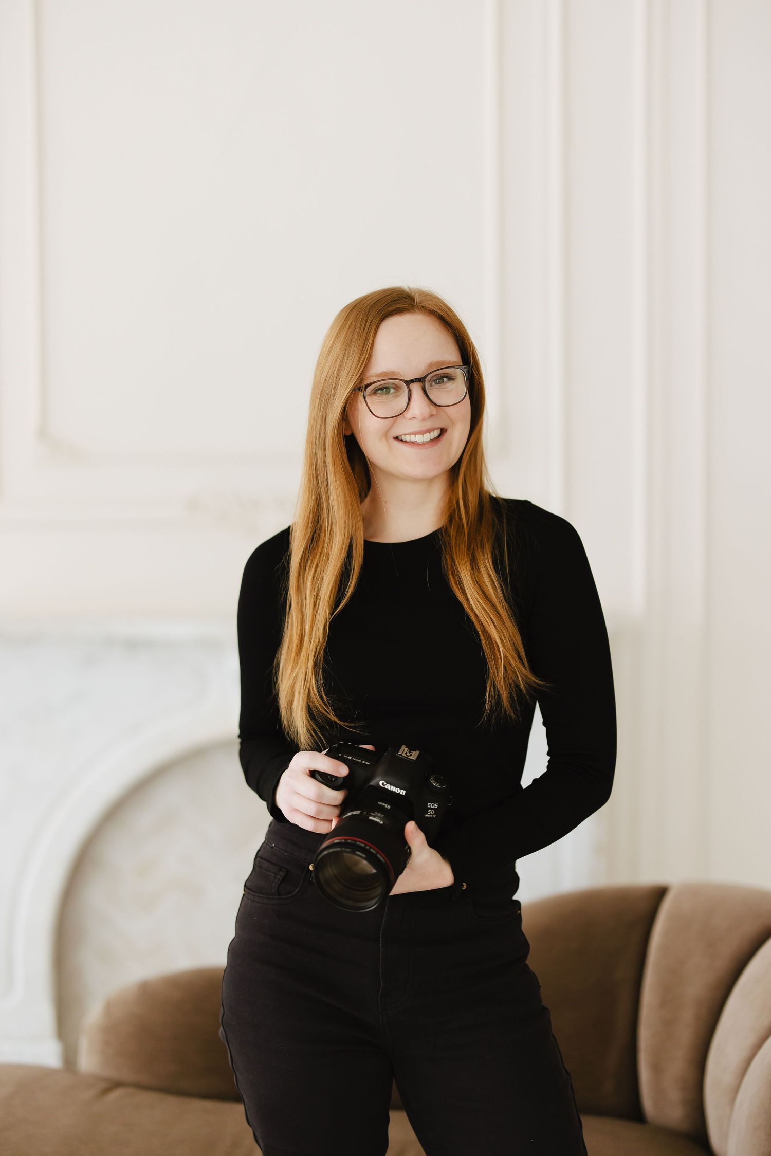 Smiling redhead woman with glasses holding a DSLR camera, wearing black outfit, posed in bright white elegant room.
