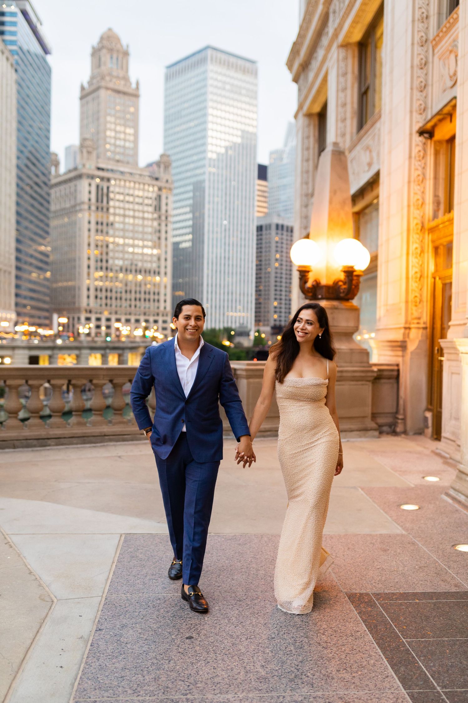 Elegant couple walks hand-in-hand past Chicago skyscrapers, man in navy suit, woman in gold gown at dusk.