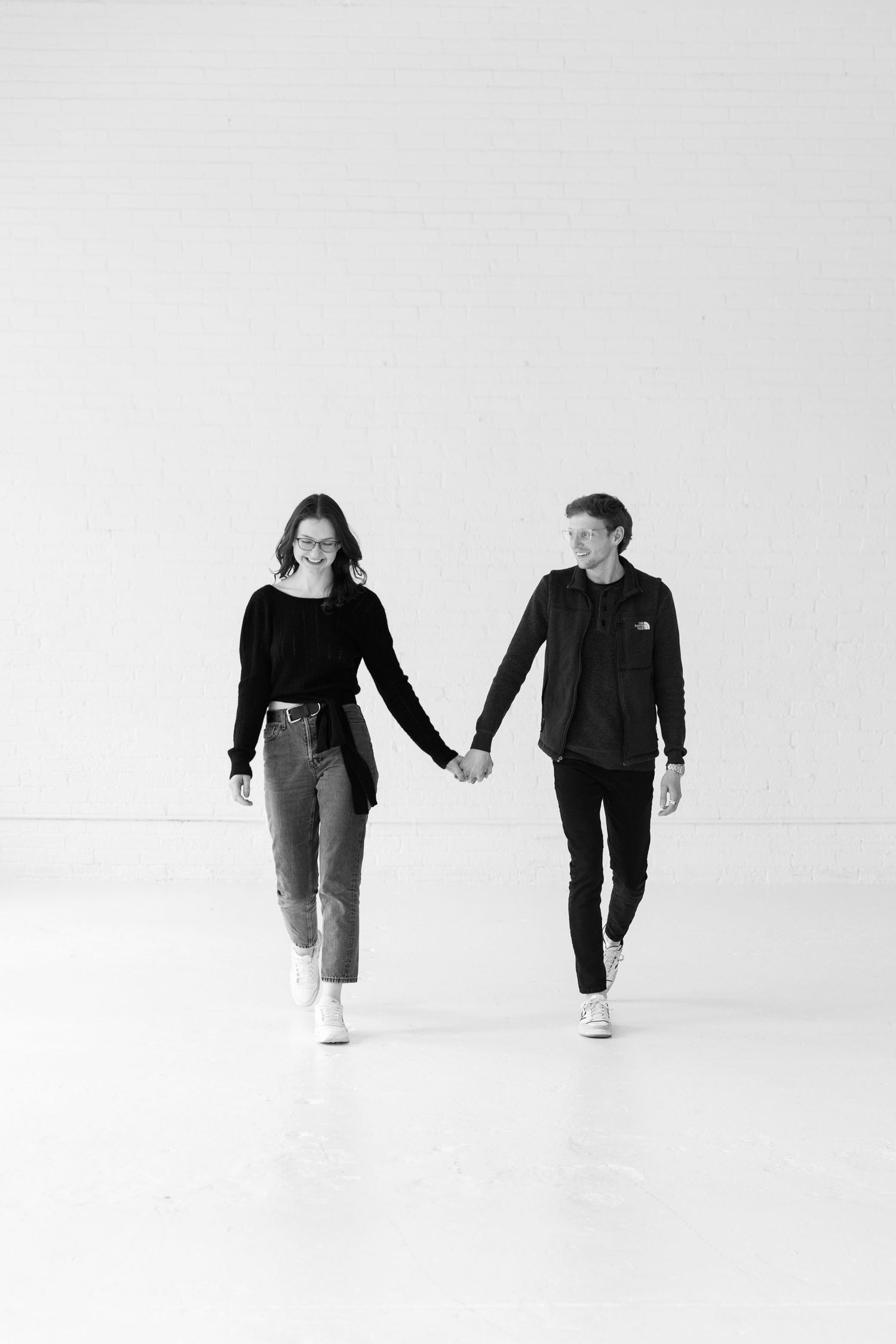 Black and white photo of a young couple holding hands while walking toward camera in a white studio.