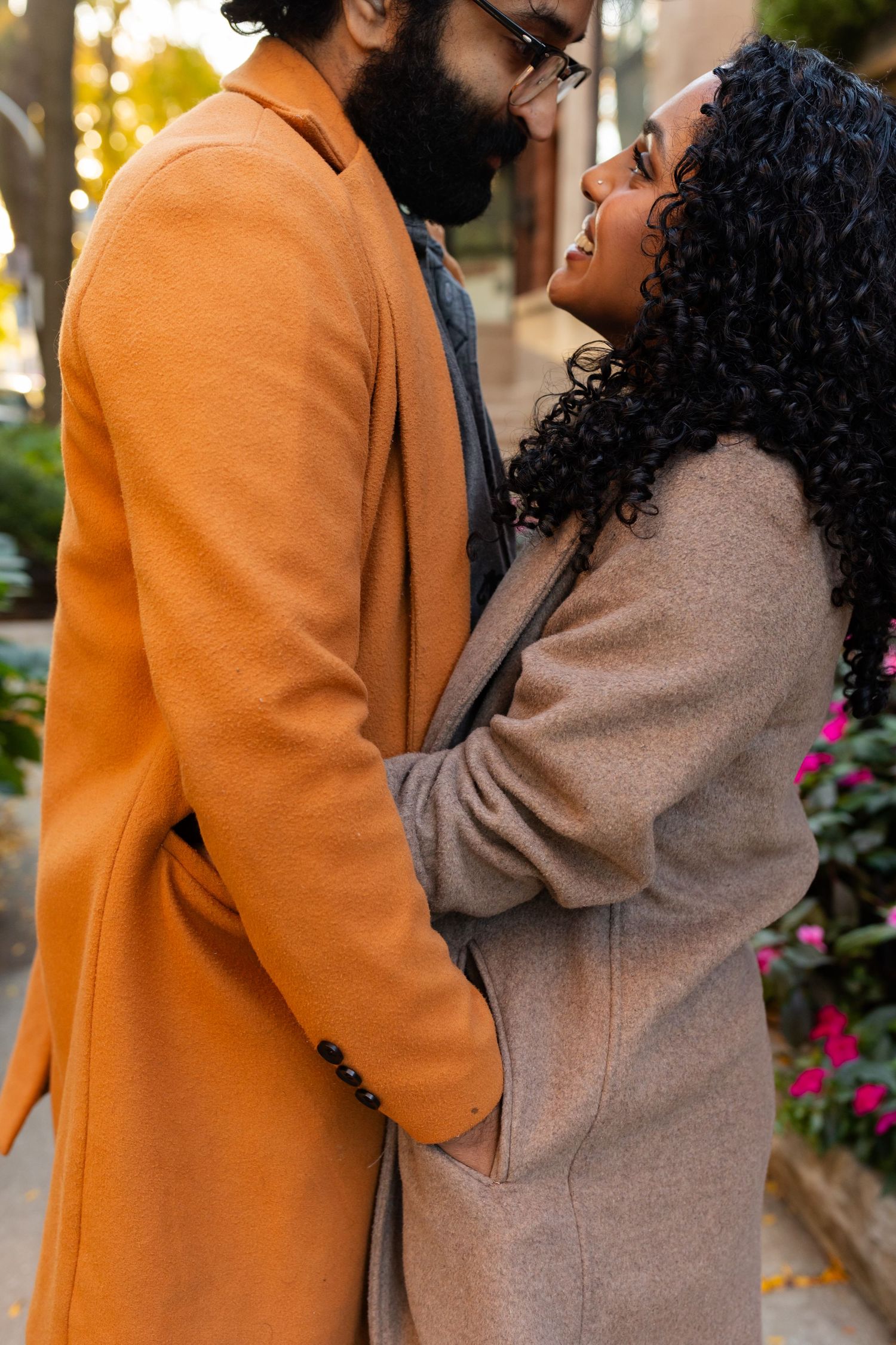 A couple embraces on a city sidewalk, with the woman looking up at her partner surrounded by flowers.