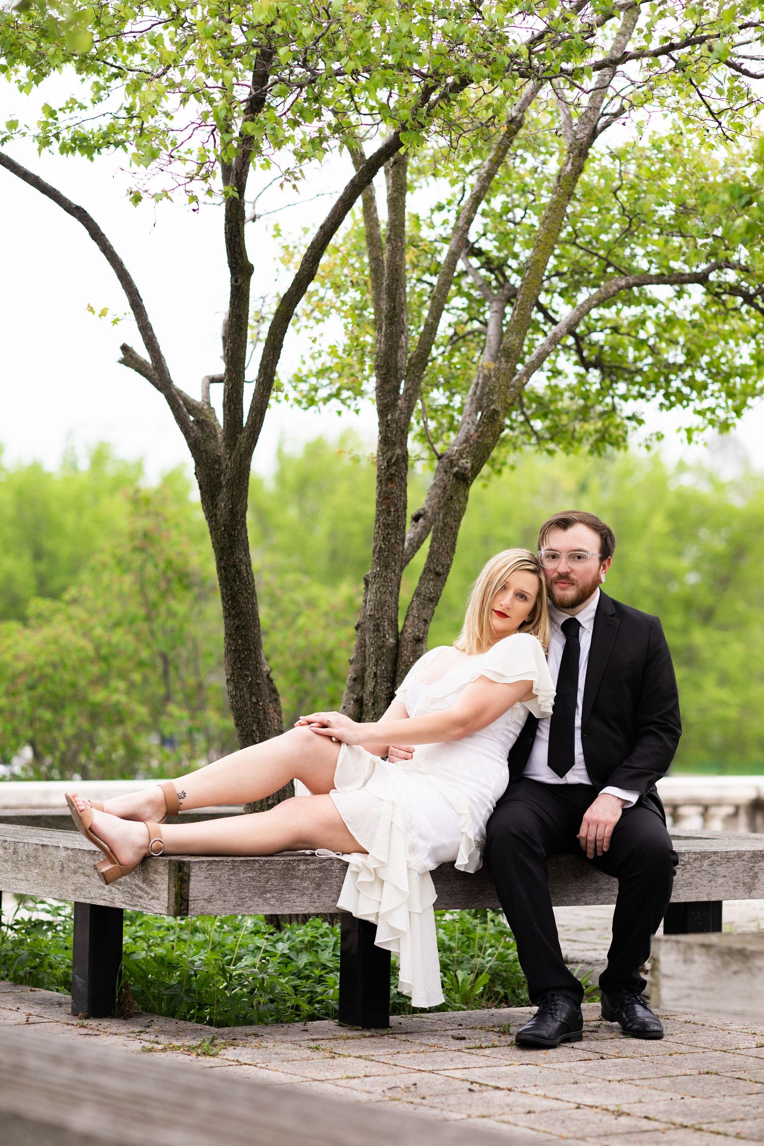 Elegant couple posing on a park bench surrounded by fresh spring greenery.
