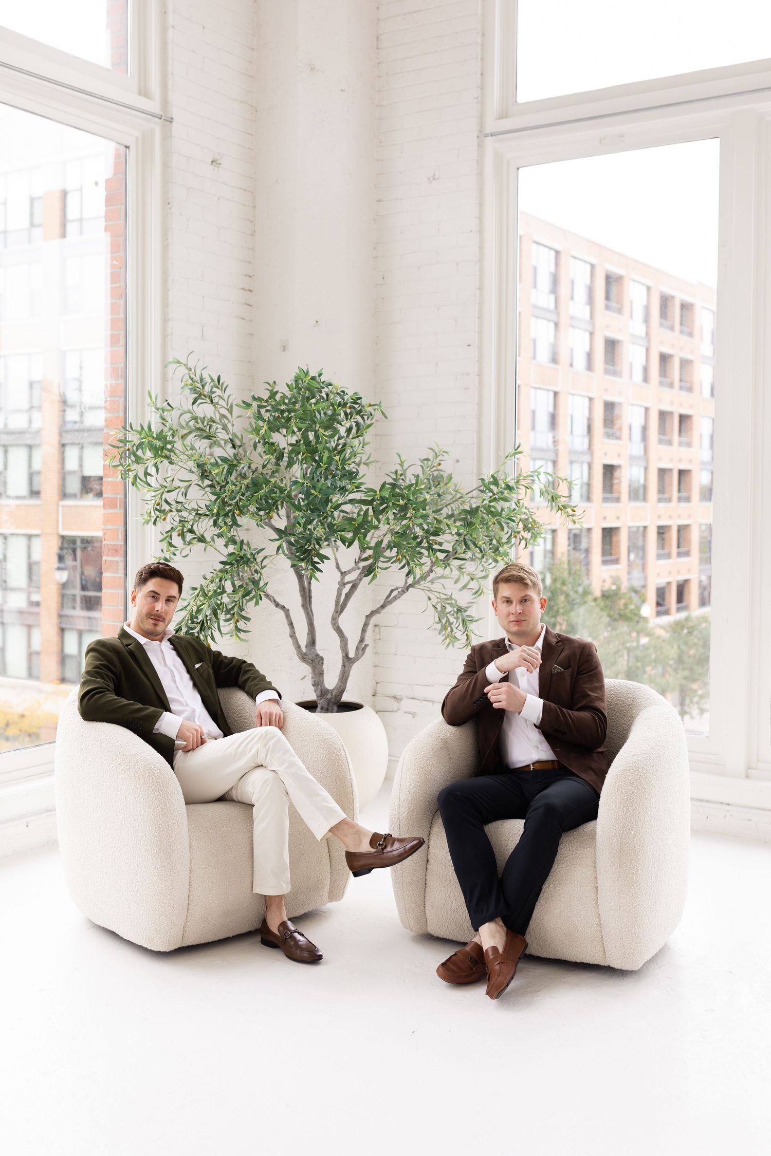 Two men in elegant suits sitting in white armchairs near large windows with city view.