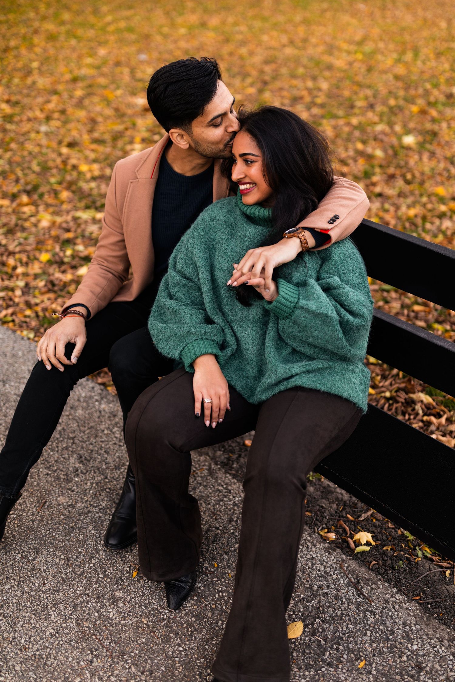Couple sitting on bench in autumn park, man kissing womans cheek as she laughs, surrounded by fallen leaves.