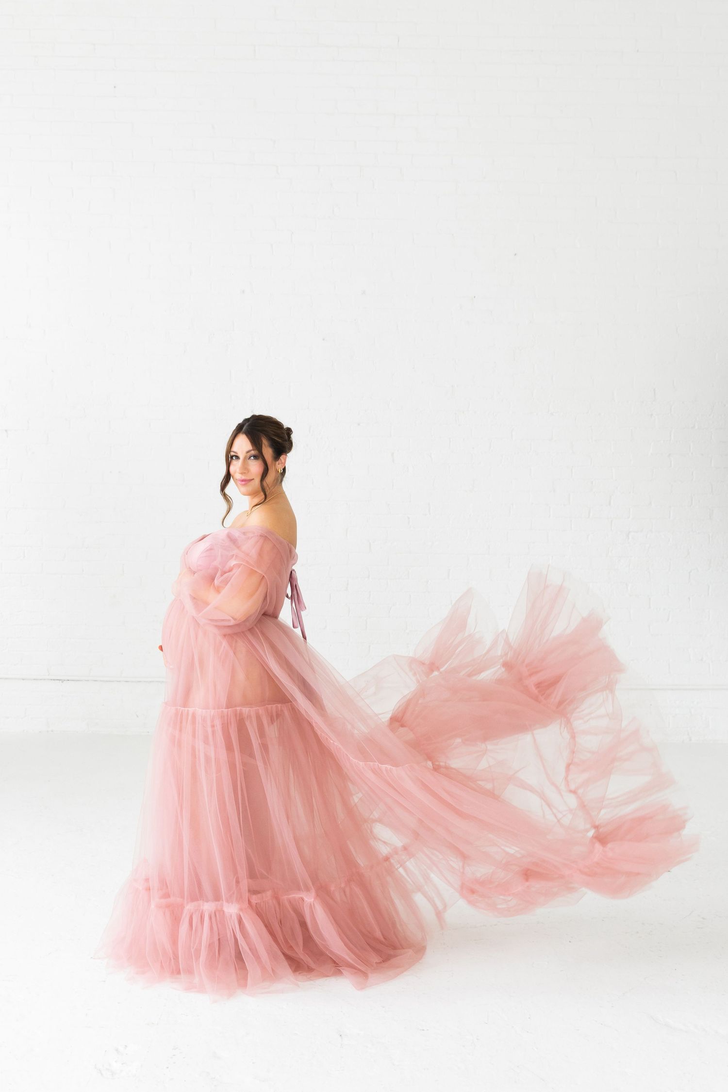 Pregnant woman in flowing pink tulle gown posing gracefully against white background.