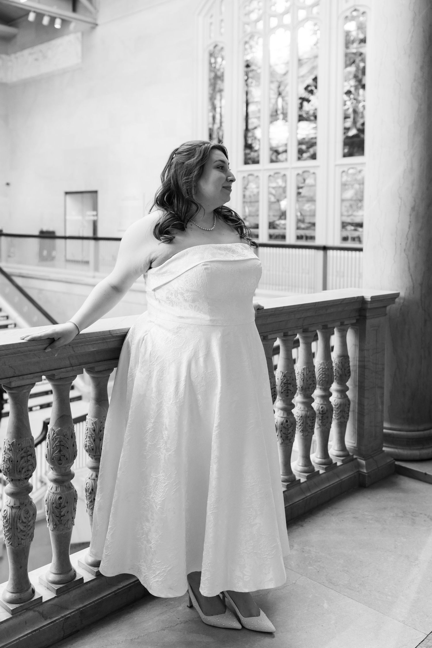 Black and white portrait of a bride in a strapless white tea-length gown standing by ornate stone balustrade.