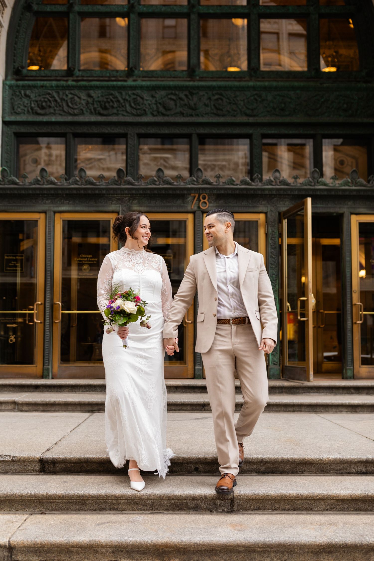 Happy bride and groom walking down steps of building 78, holding hands, bride carrying colorful bouquet.