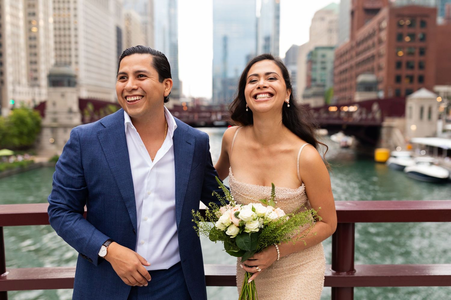 A happy couple laughs together on a bridge overlooking the Chicago River, bride holding white roses.