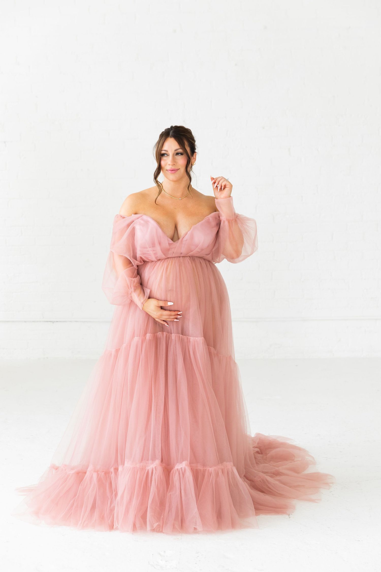 Pregnant woman in off-shoulder pink tulle maternity gown posing in bright white studio.