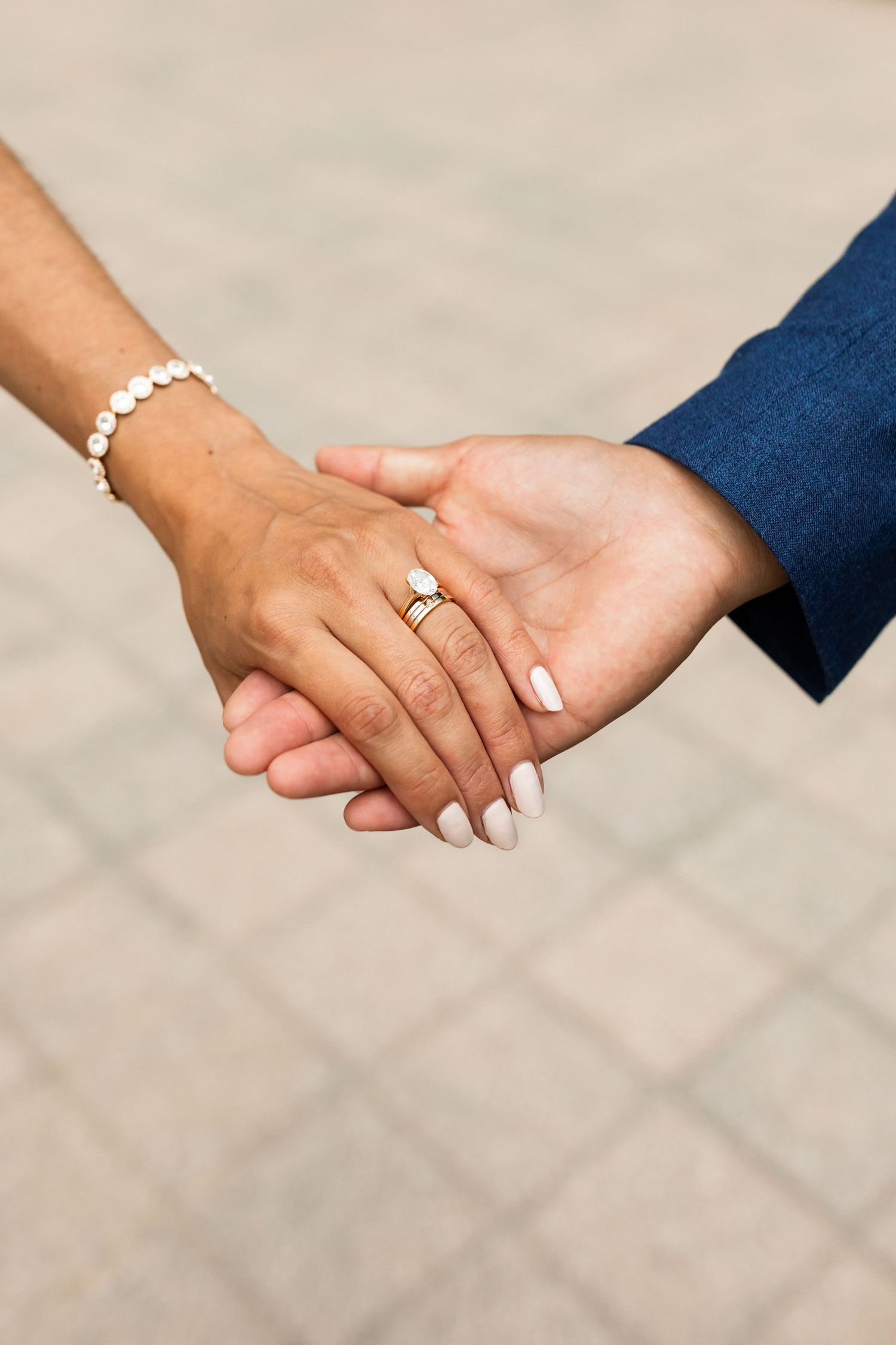 Close-up of bride and groom holding hands showing gold engagement ring and pearl bracelet outdoors.