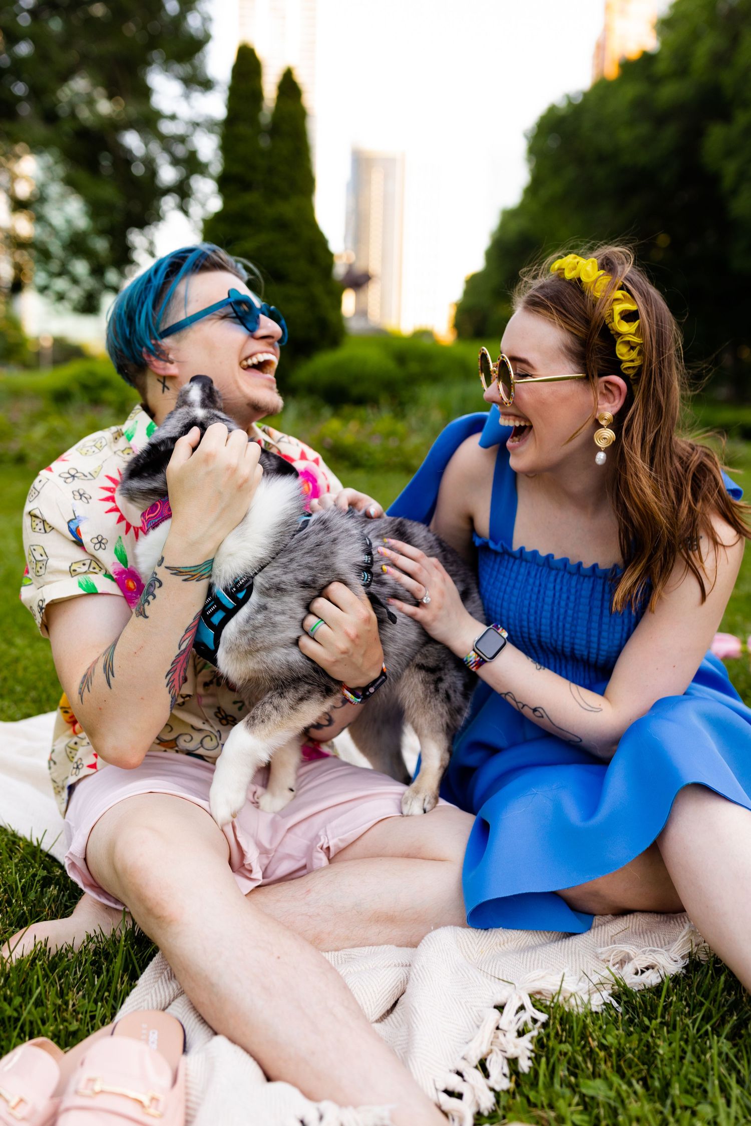 Laughing couple with blue-haired partner playing with a husky puppy on park grass.