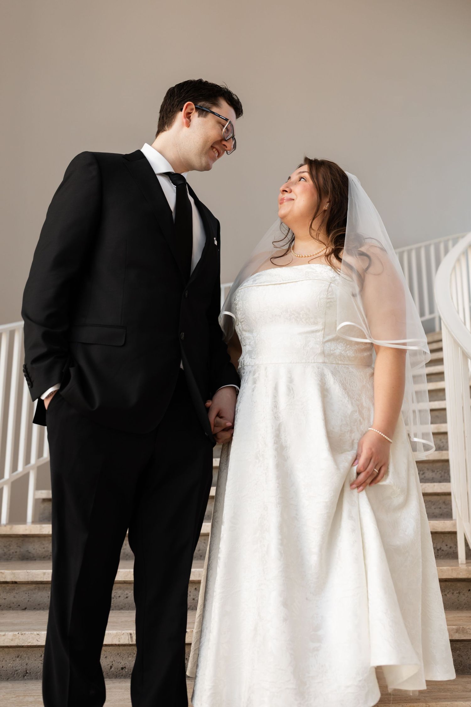 Bride and groom holding hands and smiling at each other on marble staircase in elegant venue.