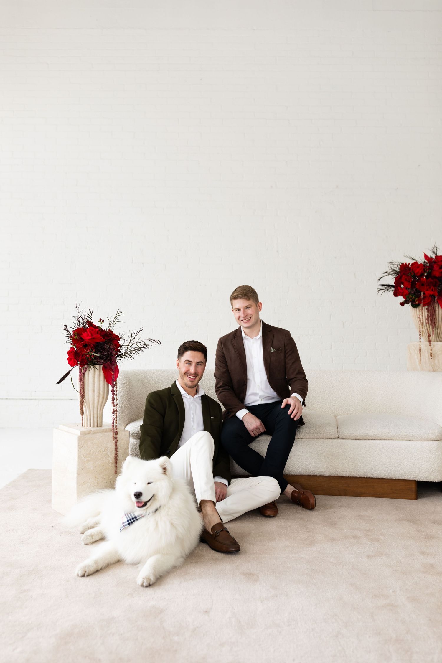 Two grooms in brown and green blazers pose with white Samoyed dog beside white sofa with red floral arrangements.