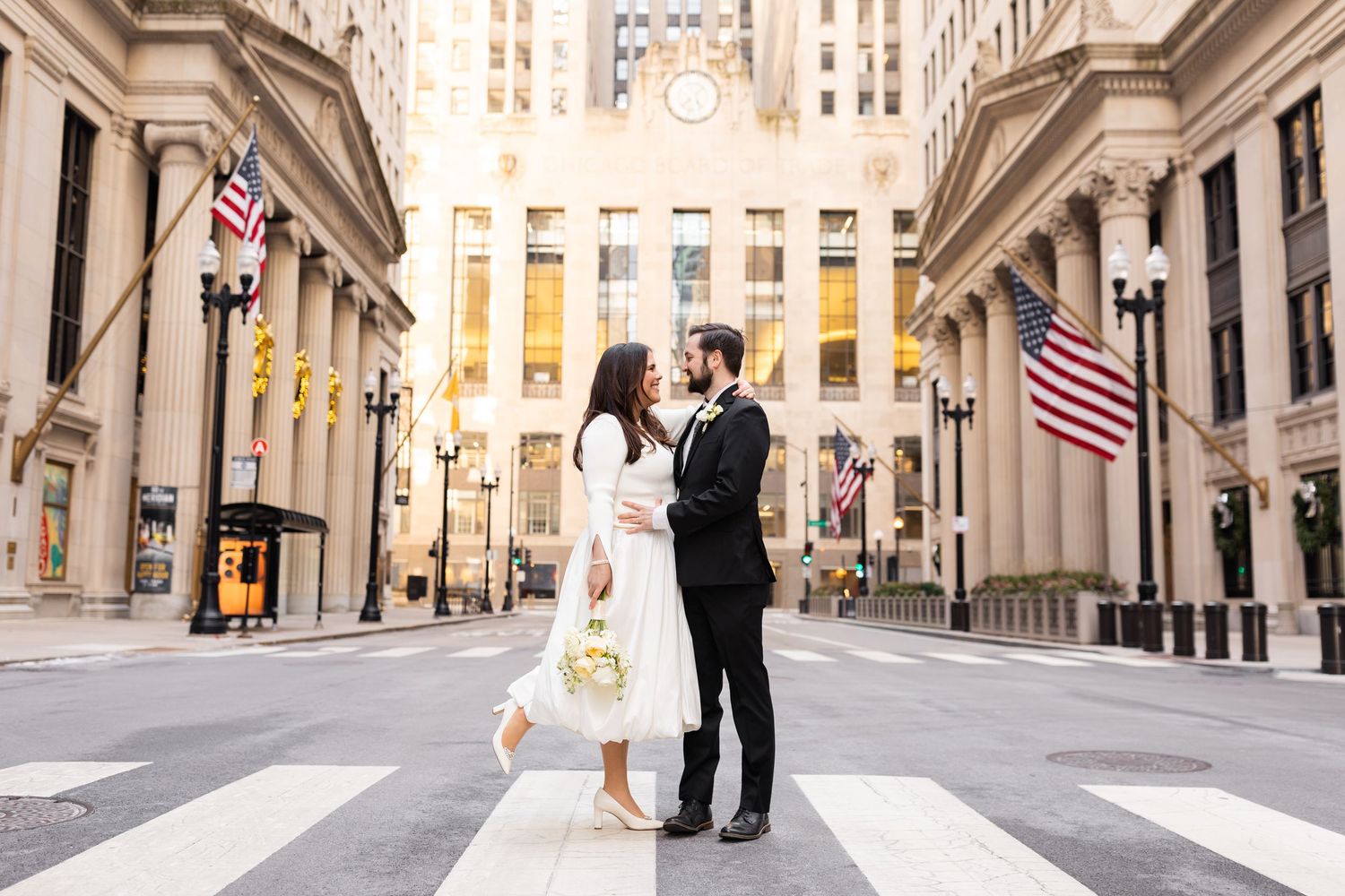 Bride and groom standing on crosswalk in front of Chicago Board of Trade building with American flags.