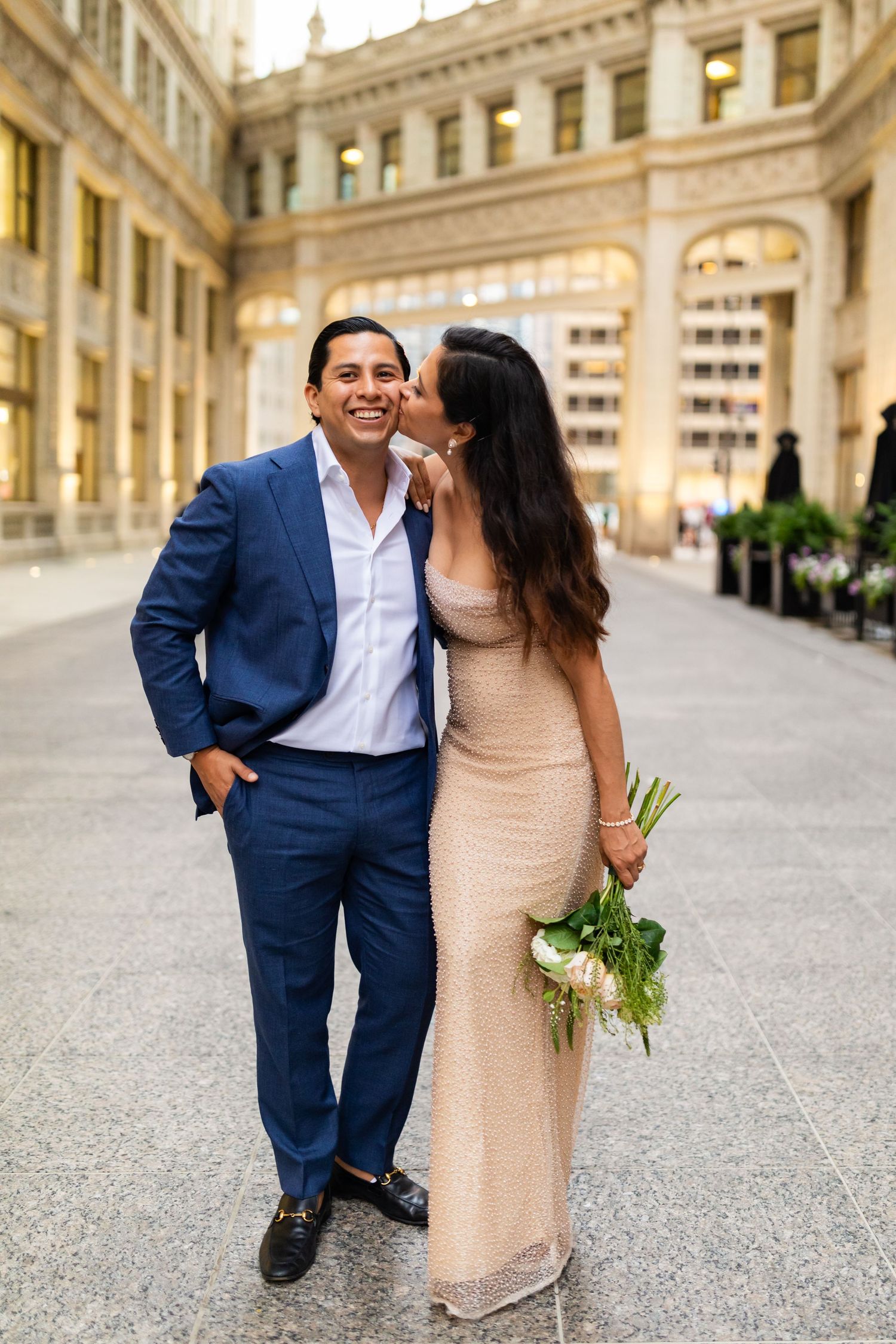 Woman kissing man's cheek as he smiles holding bouquet in elegant Chicago Wrigley Building courtyard.