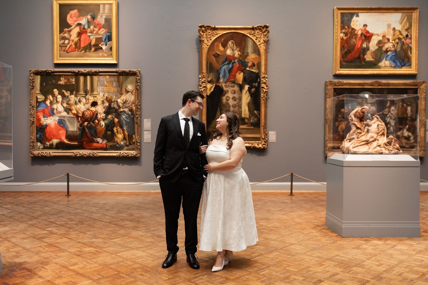 Bride in white dress and groom in tuxedo share joyful moment surrounded by classical paintings.