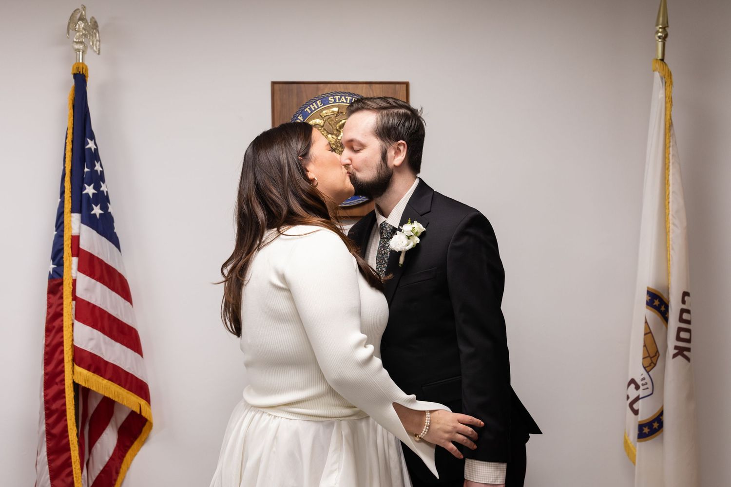 Newlyweds sharing their first kiss at a city hall wedding ceremony beside American and state flags.