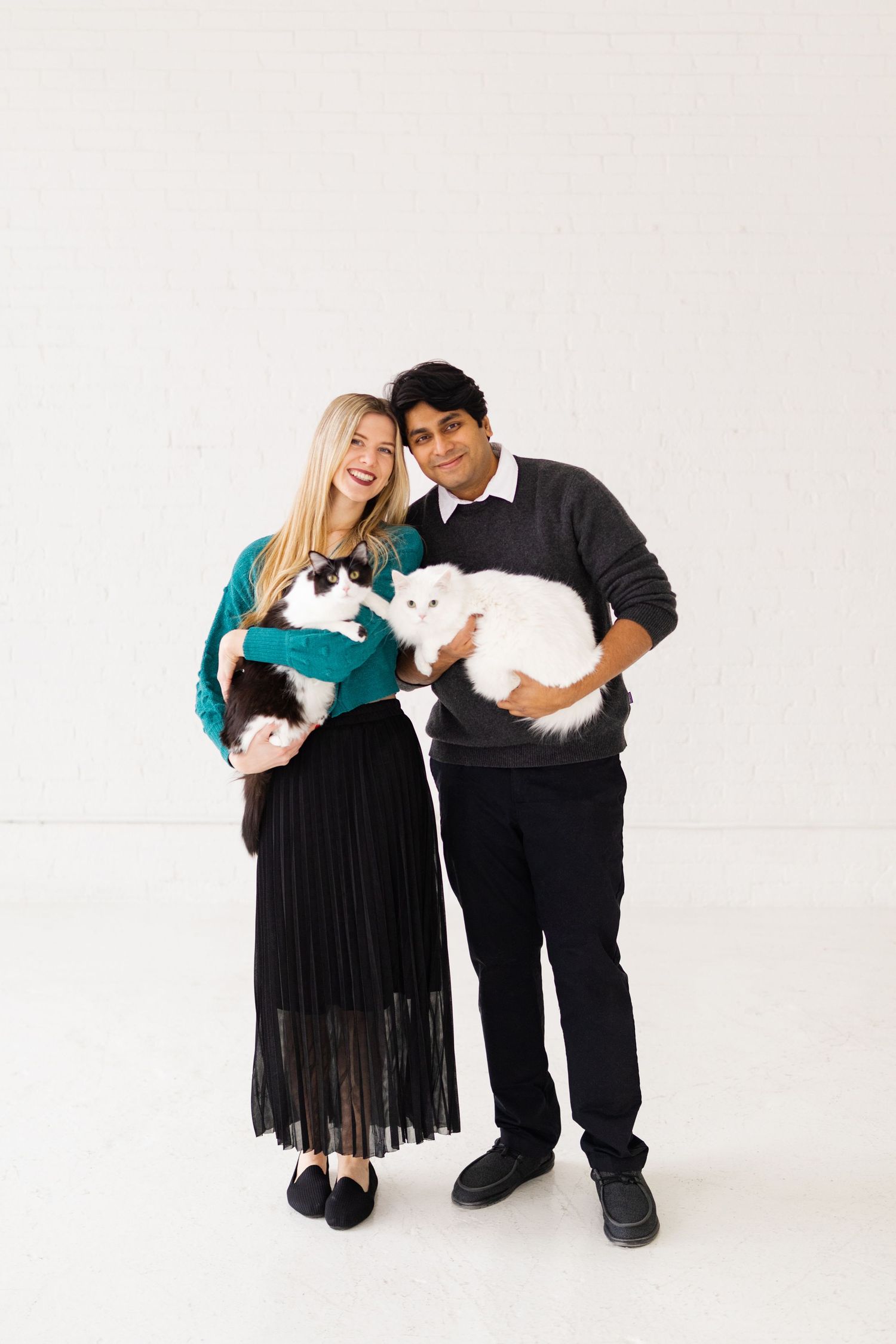 Happy couple posing with black-and-white and white fluffy cats in a bright studio.