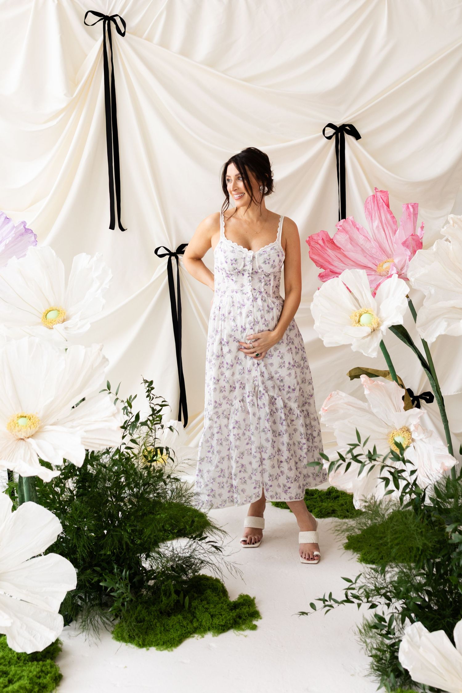Pregnant model in floral sundress stands among giant white paper flowers and lush green plants.