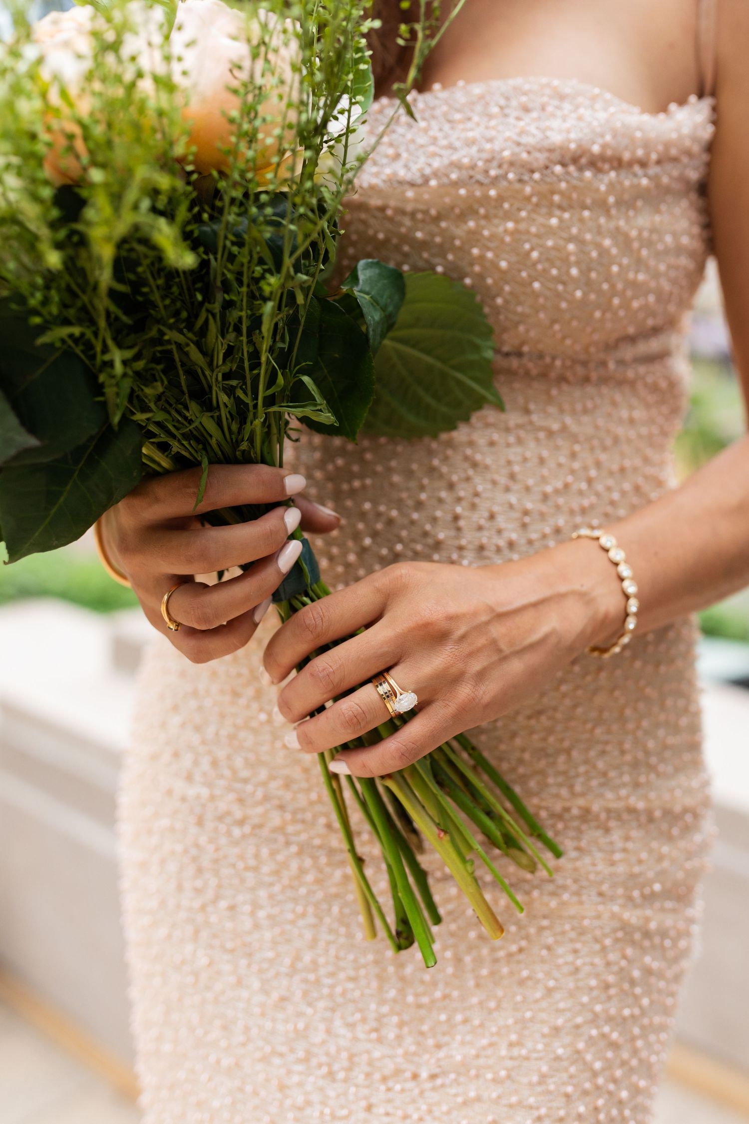 Close-up of bride holding floral bouquet wearing pearl bracelet and gold wedding rings in sparkly dress.