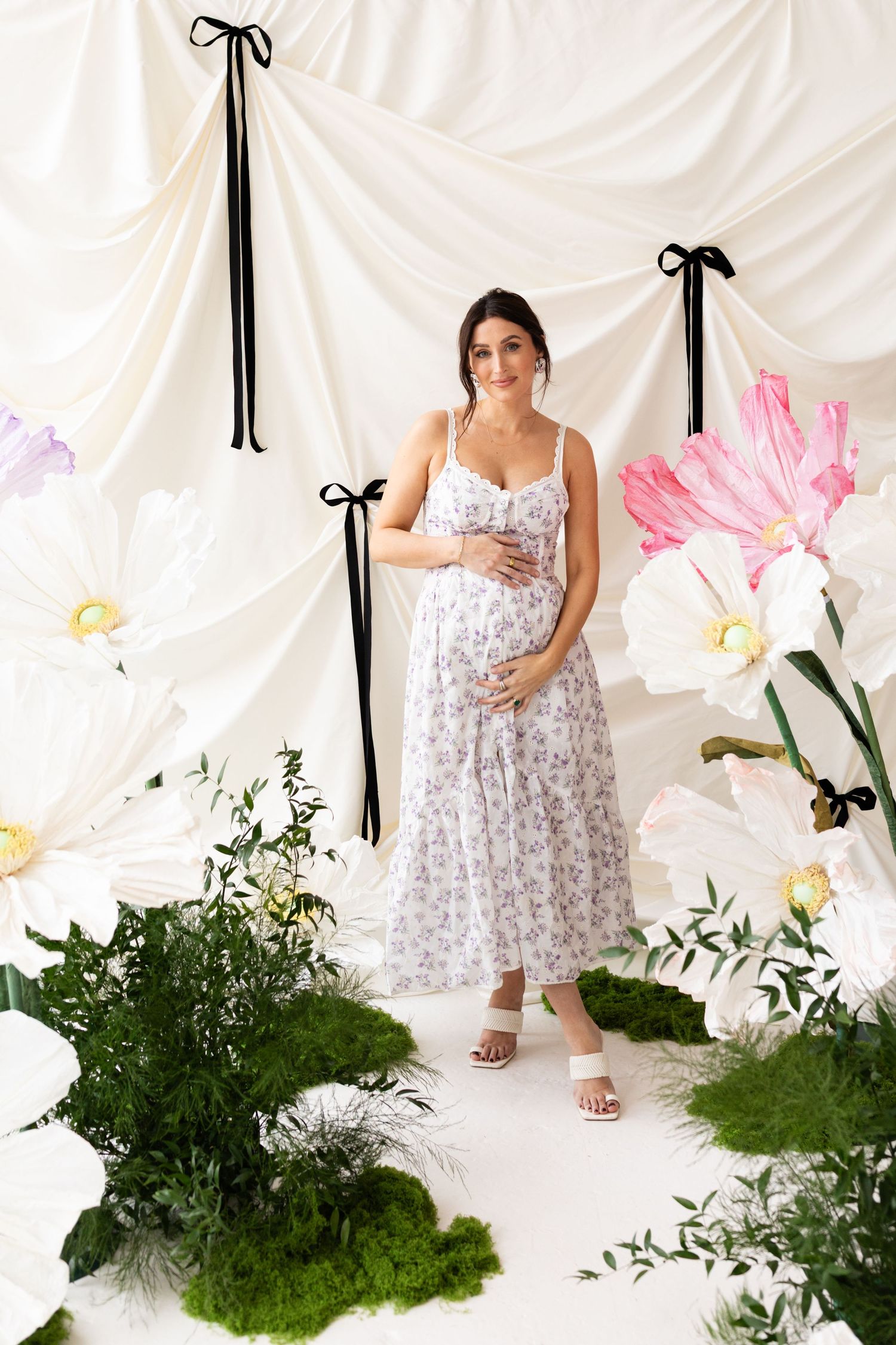 Smiling pregnant woman in white floral midi dress posing among oversized flowers and green plants.