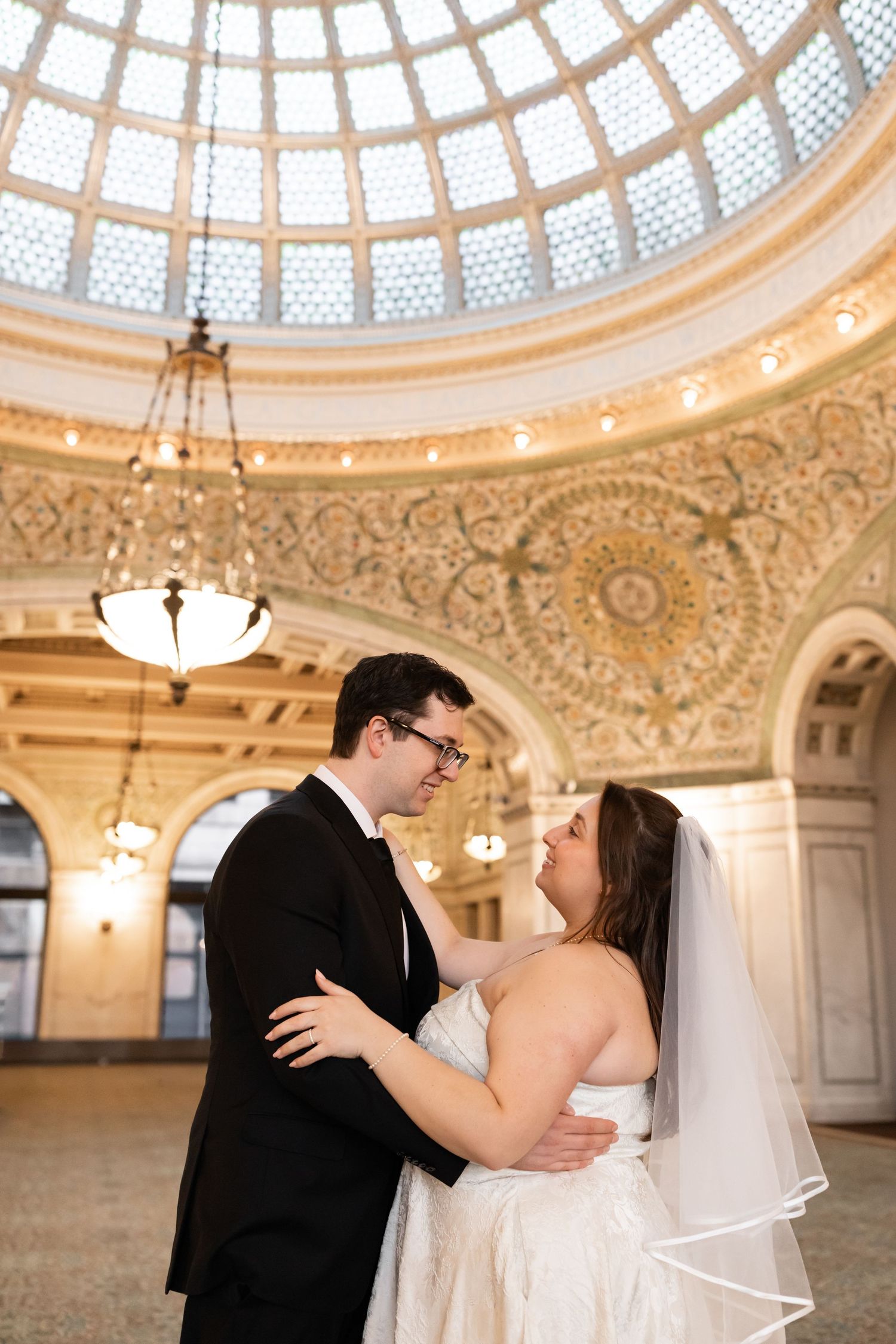 Bride and groom embrace under ornate domed ceiling with stained glass at Chicago Cultural Center.