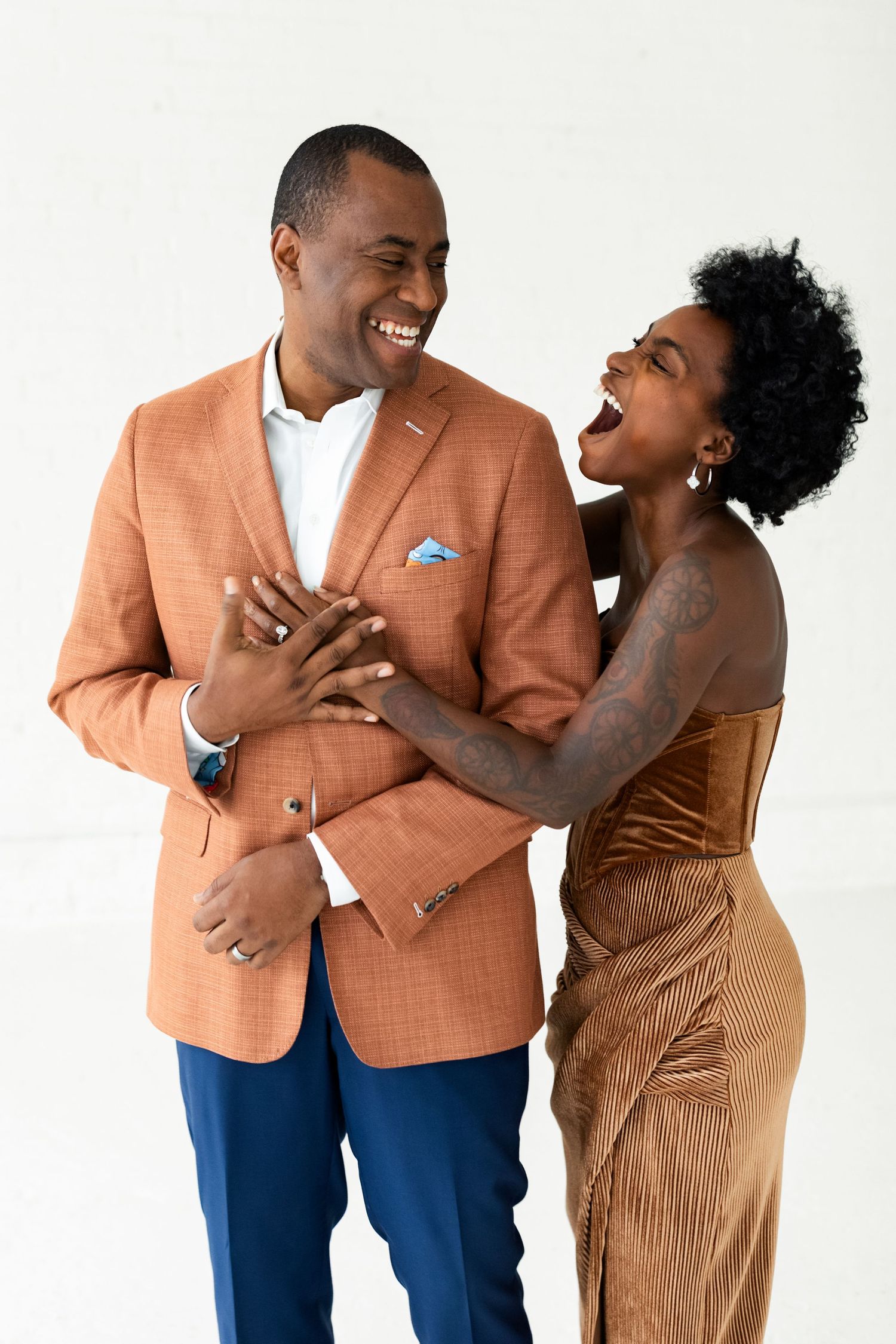A joyful couple laughing together, the woman in a gold dress embracing the man in an orange blazer.