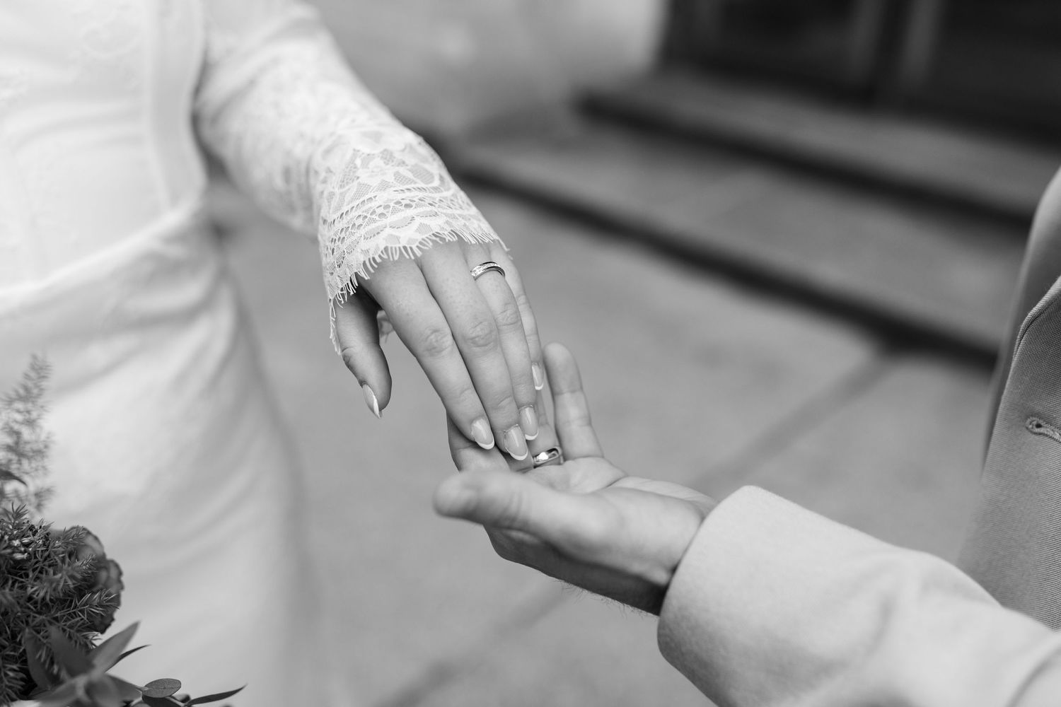 Black and white close-up of bride and groom holding hands, showcasing wedding ring on bride's finger.