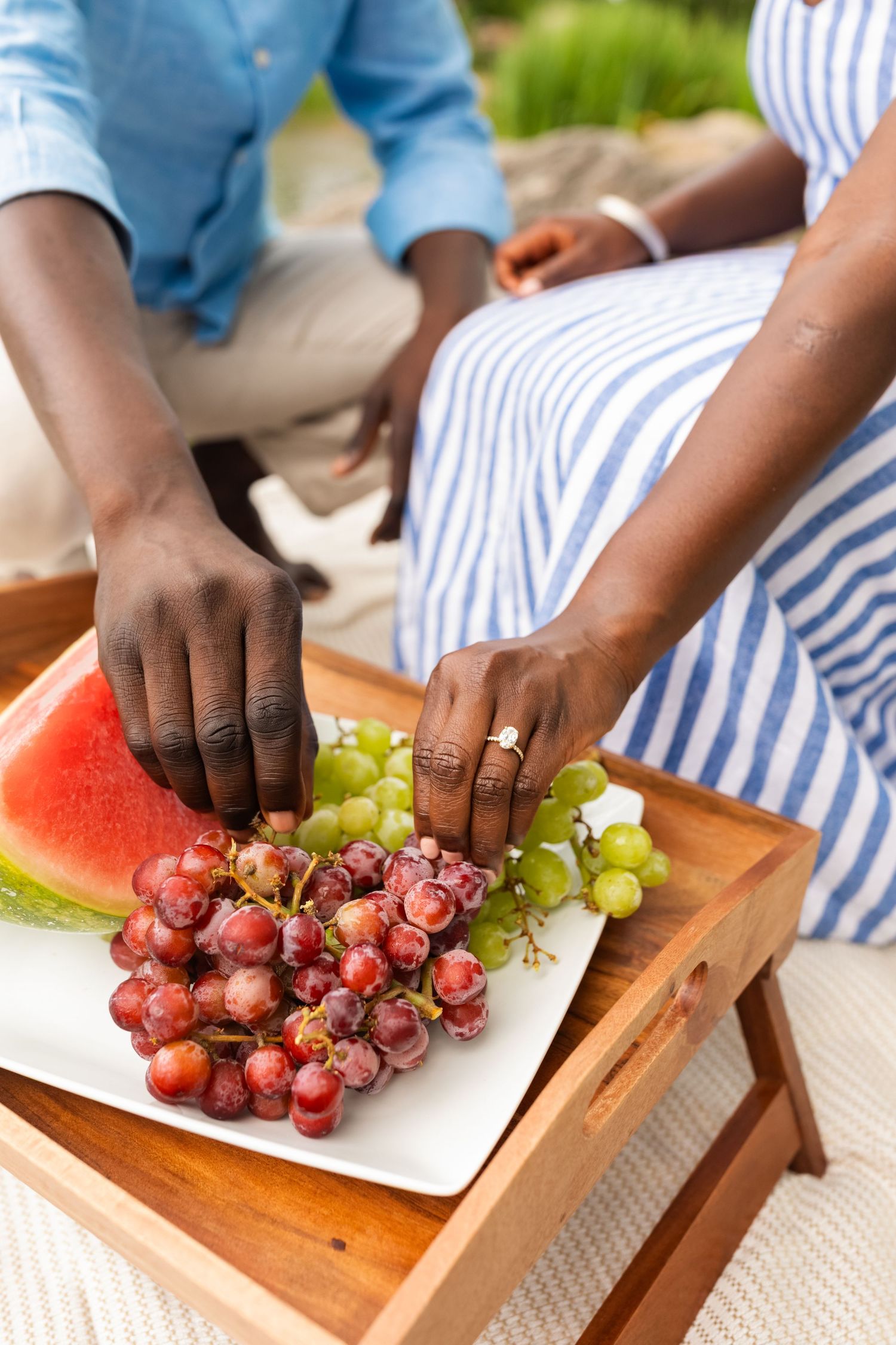 Couple enjoying a picnic with fresh grapes and watermelon on a wooden tray.