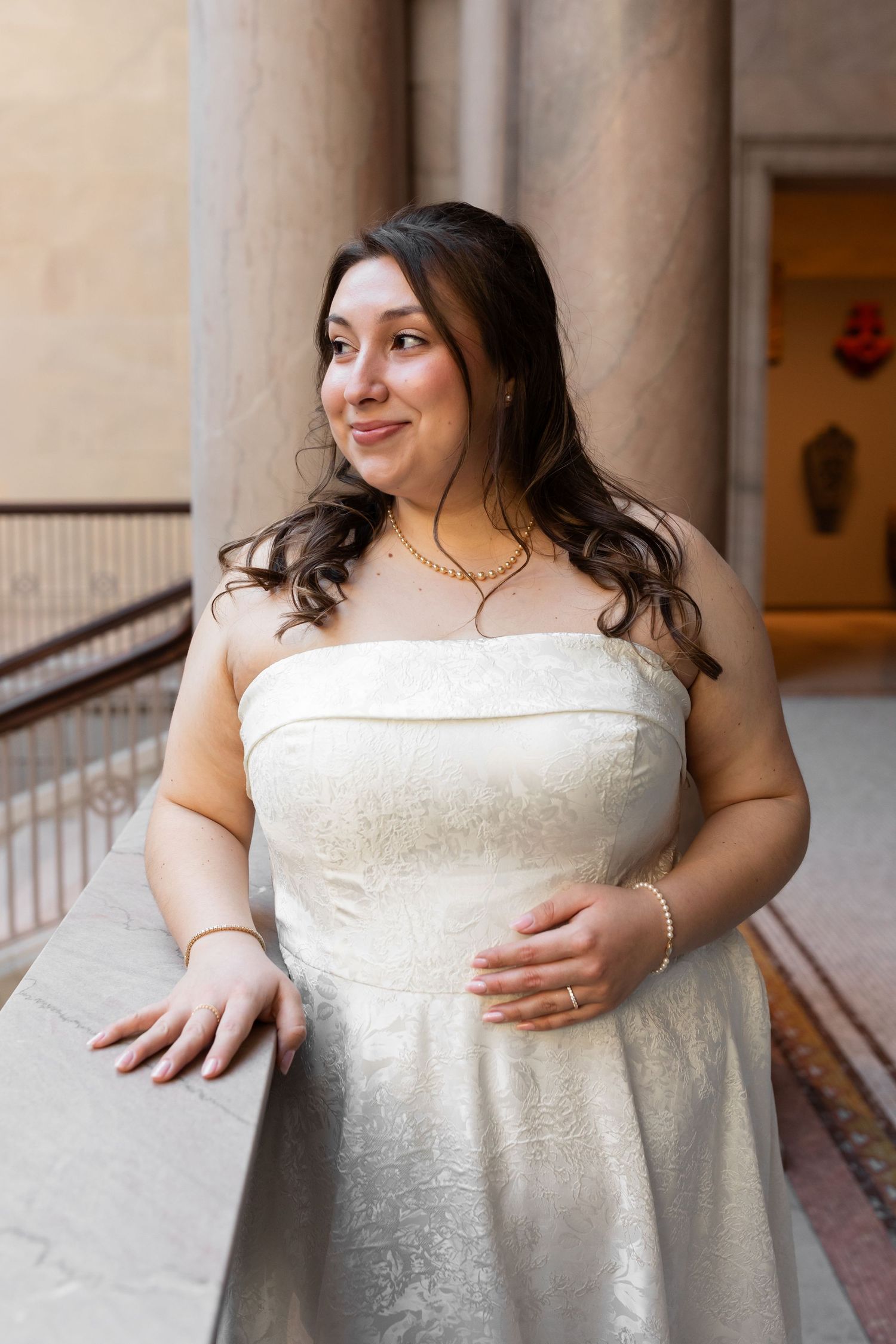 Bride in strapless ivory lace gown smiling warmly while leaning on a stone railing indoors.