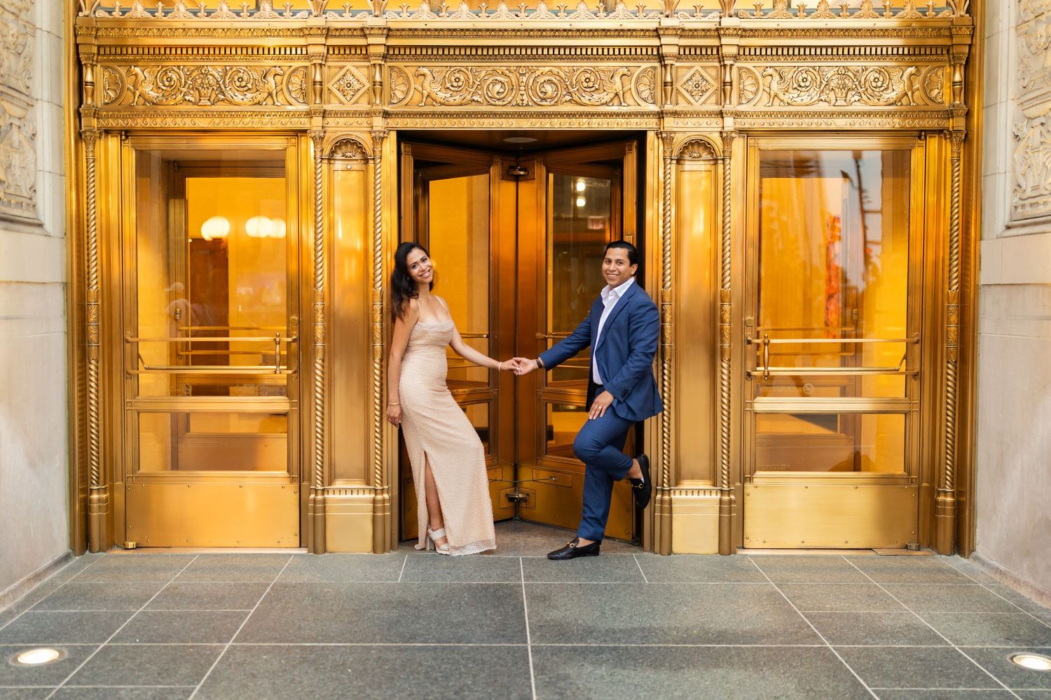 Engaged couple holding hands before ornate gold revolving doors of elegant city building at dusk.
