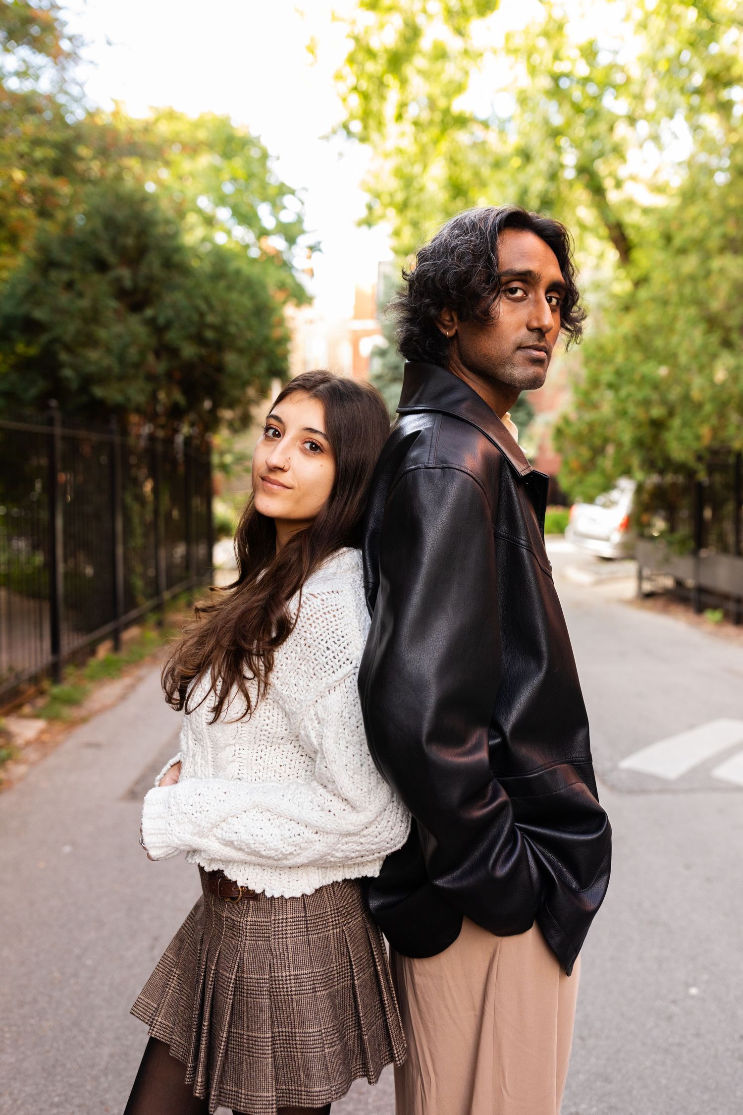 Interracial couple posing back-to-back on a tree-lined urban street, woman in white sweater and plaid skirt, man in black leather jacket.