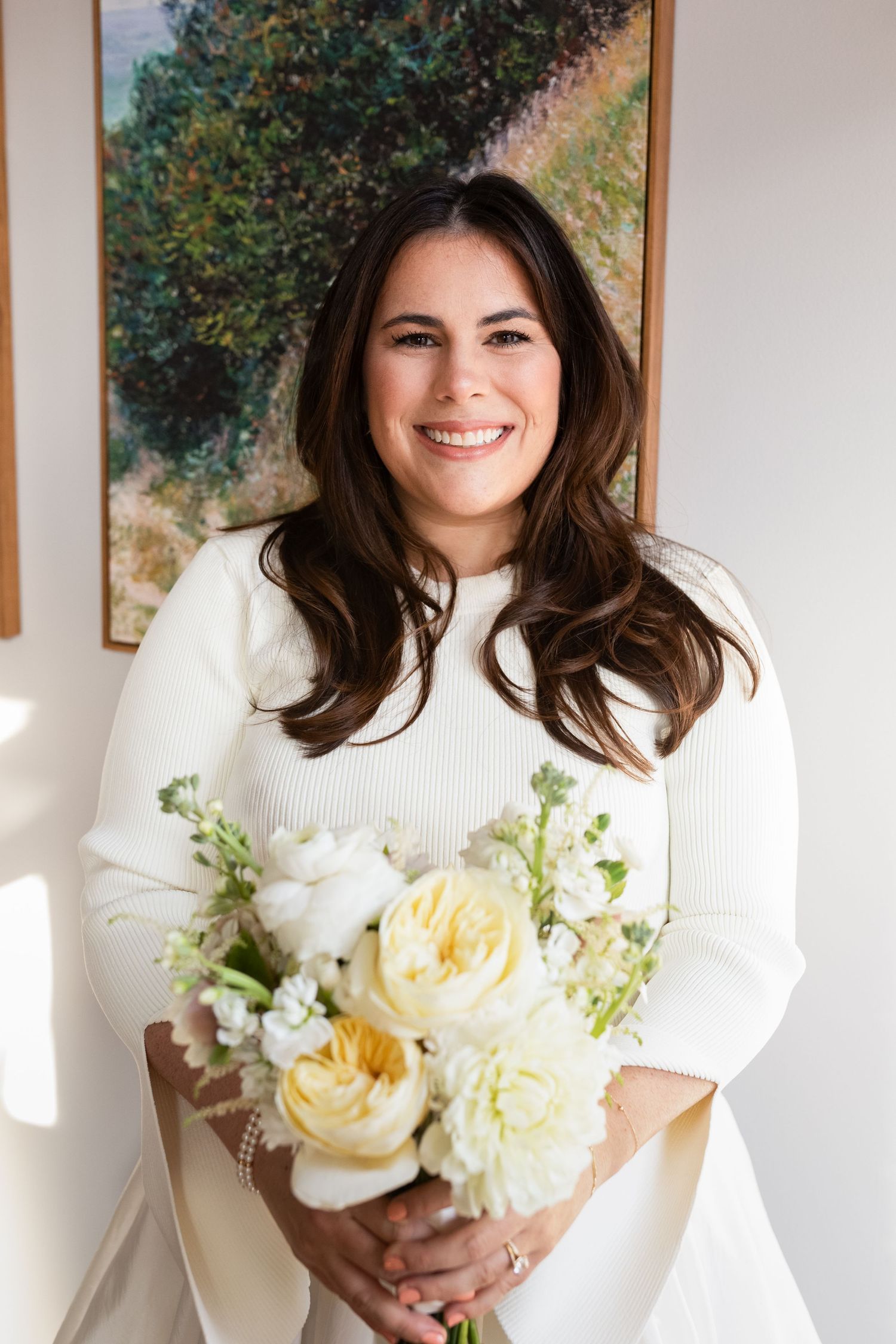Radiant bride with long dark hair smiles holding cream and yellow roses wedding bouquet.