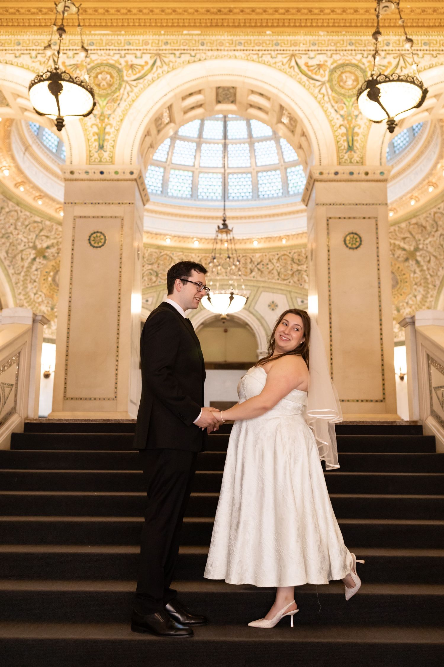 Bride and groom hold hands on ornate staircase beneath glowing stained glass dome.