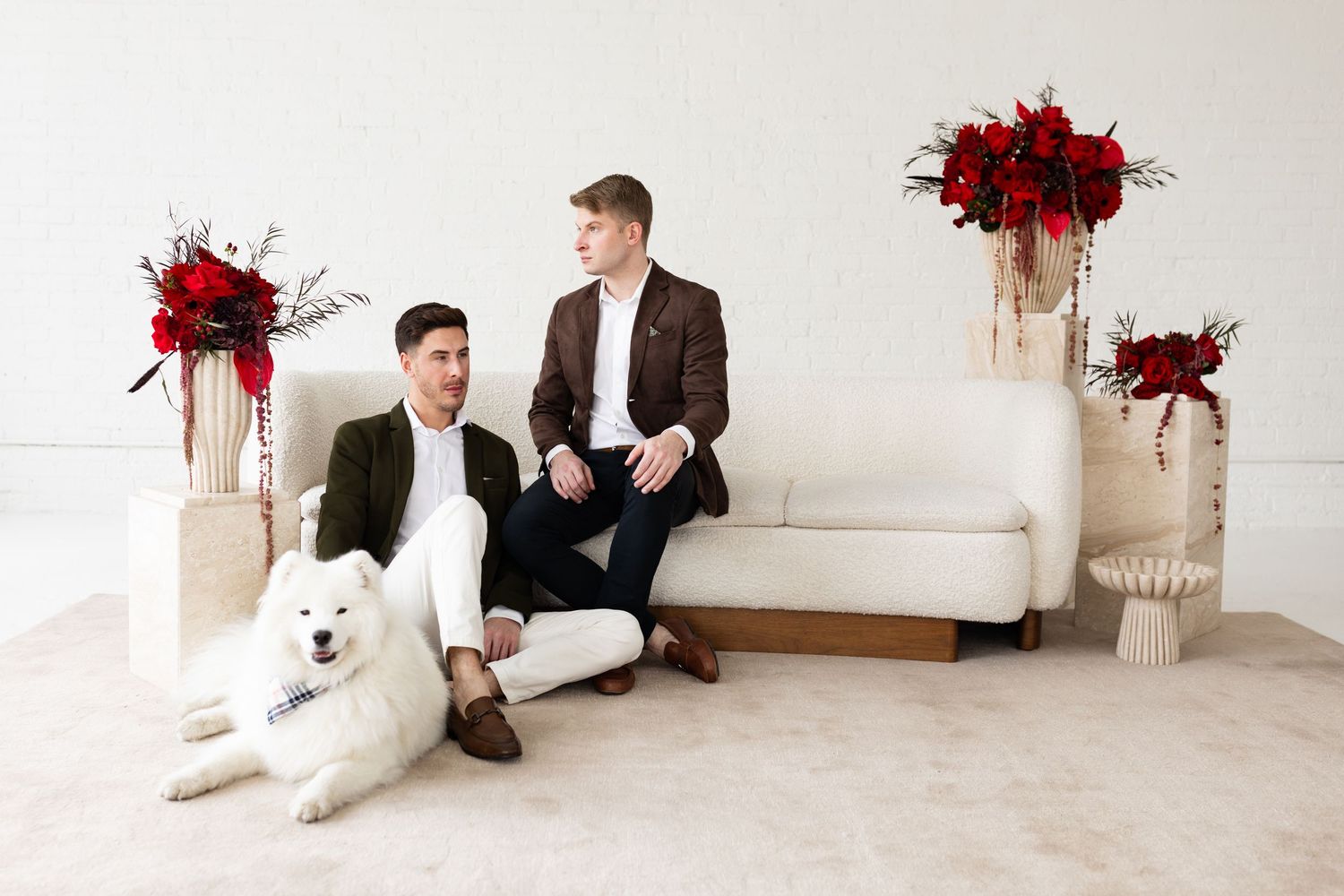 Two grooms sit with their white Samoyed dog on a cream sofa surrounded by red floral arrangements.