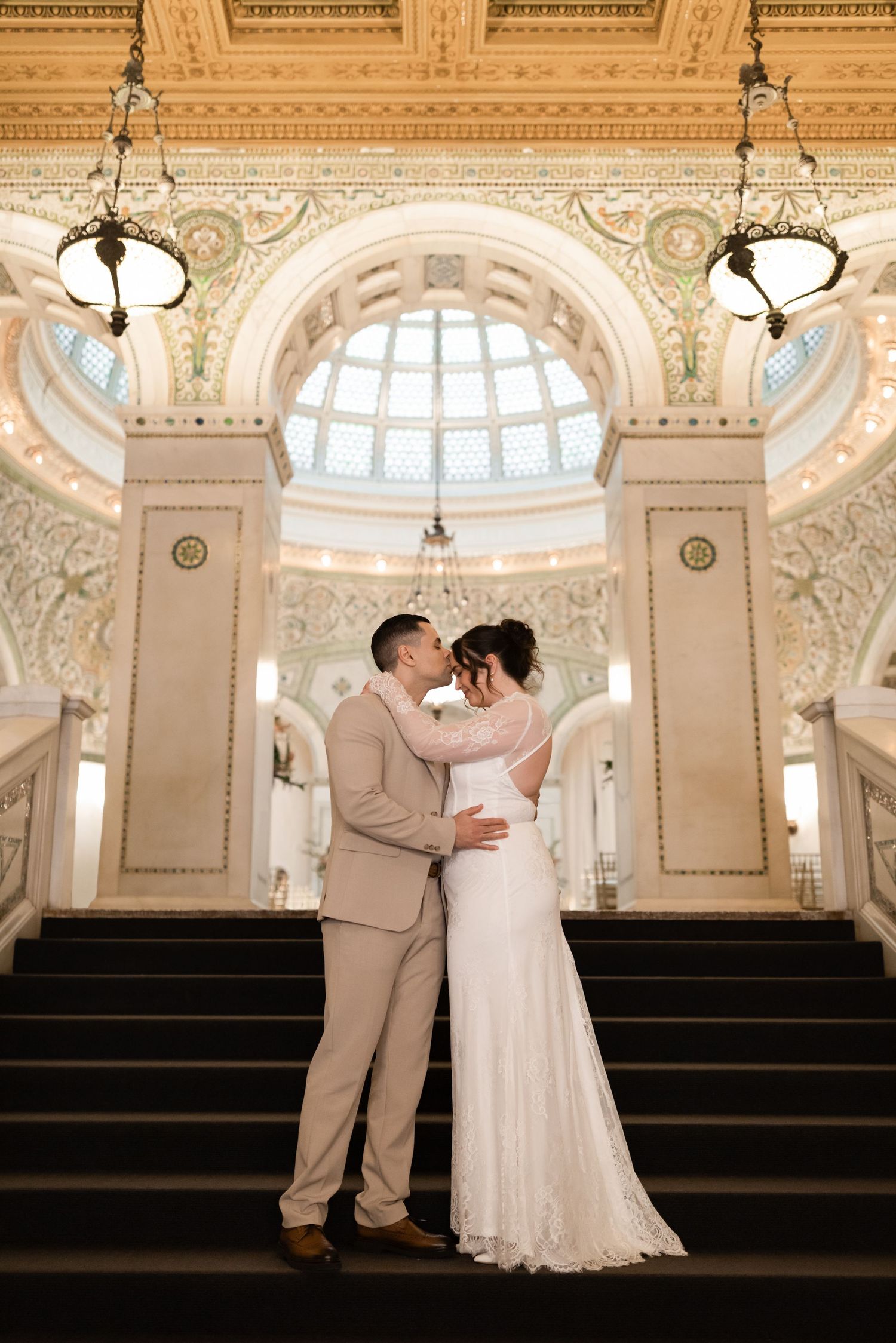 Bride and groom sharing romantic kiss on grand staircase beneath stunning mosaic dome ceiling.