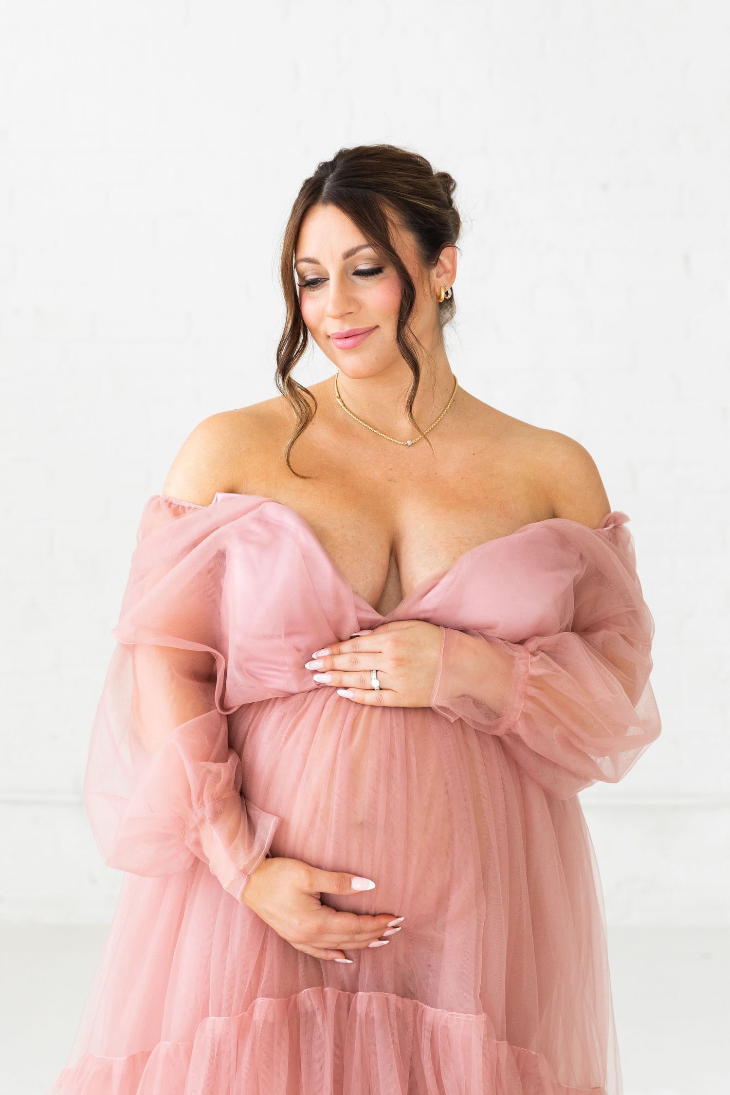 Pregnant woman in pink tulle off-shoulder gown cradling baby bump in white studio.