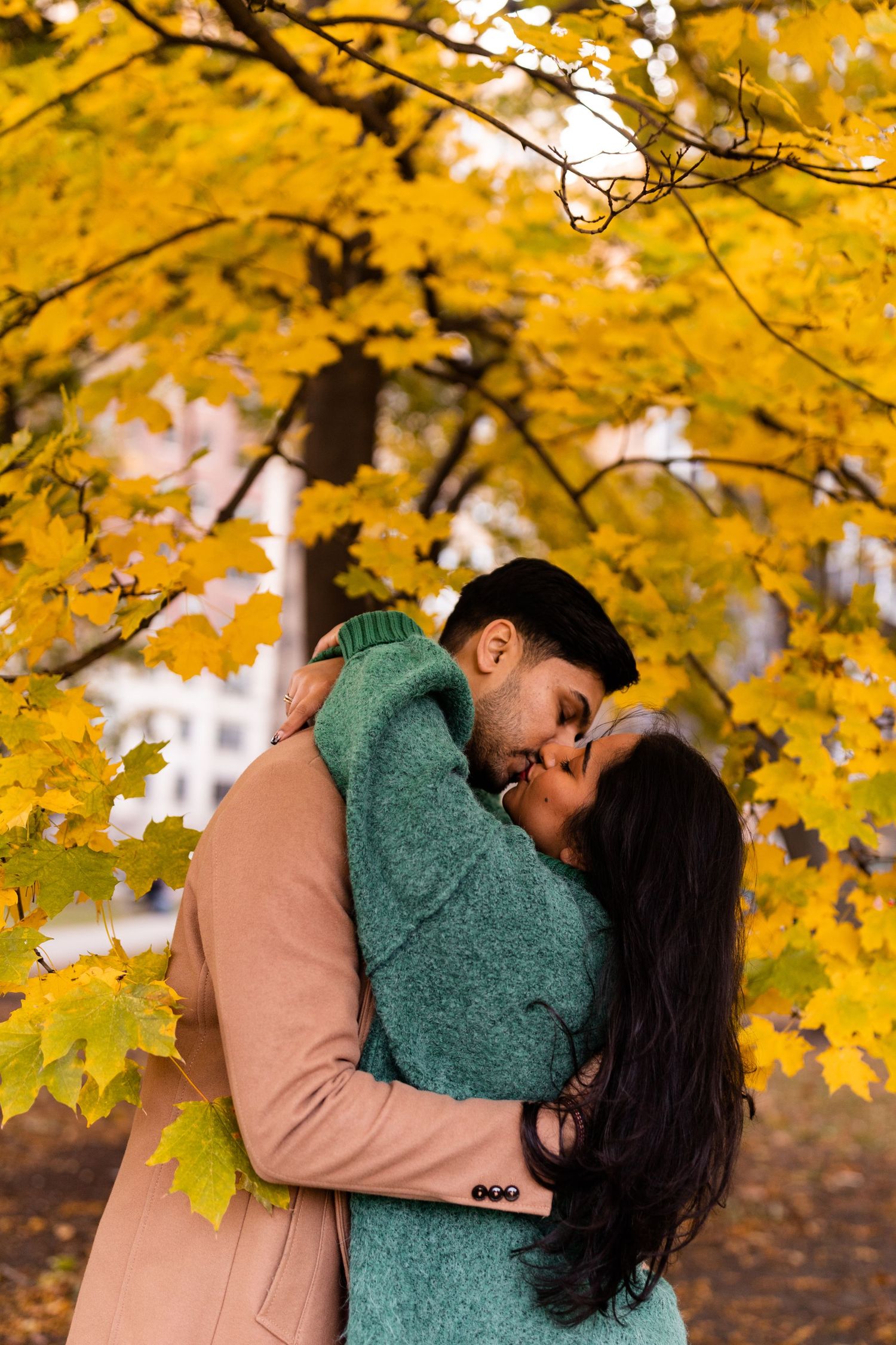 Couple sharing a kiss under golden autumn maple tree, man in camel coat embracing woman in teal oversized sweater.