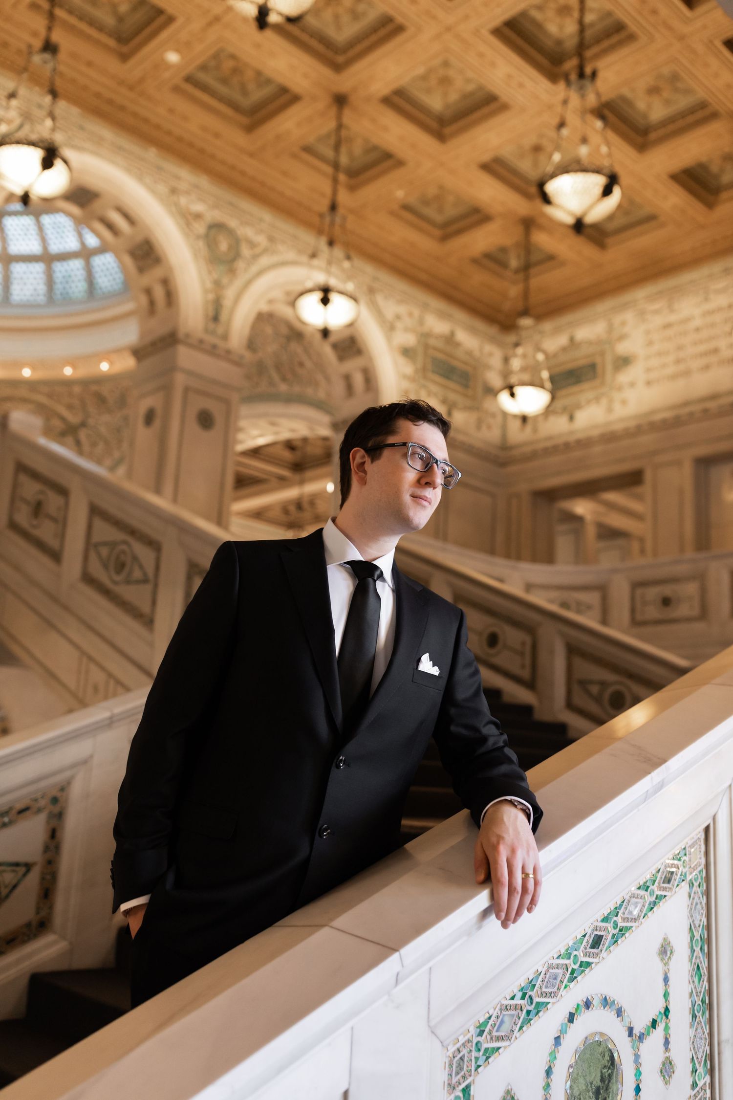 Groom in a black suit leaning on an ornate marble staircase railing inside a grand historic building.