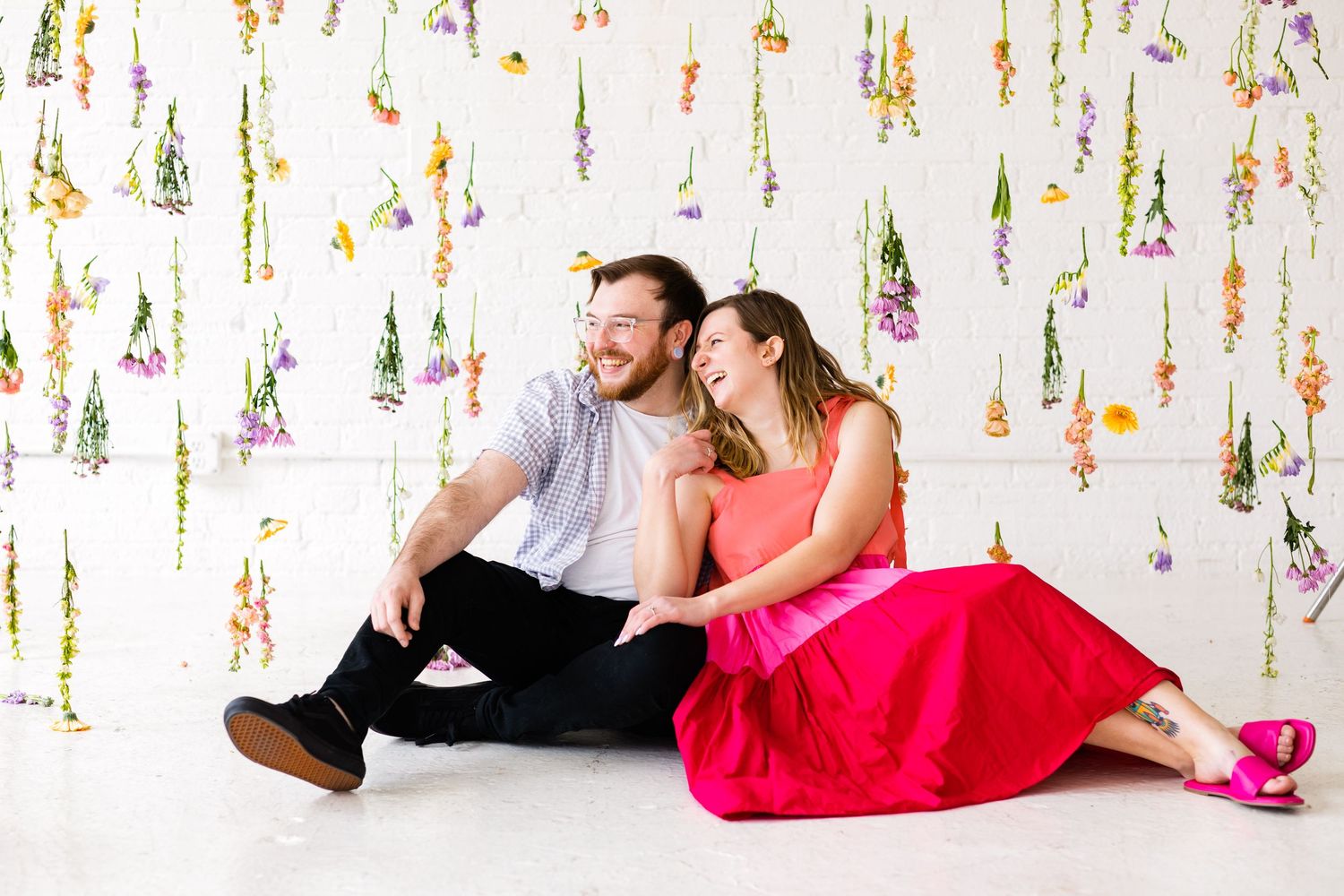Smiling couple sitting together on floor with decorative hanging wildflowers in white studio.