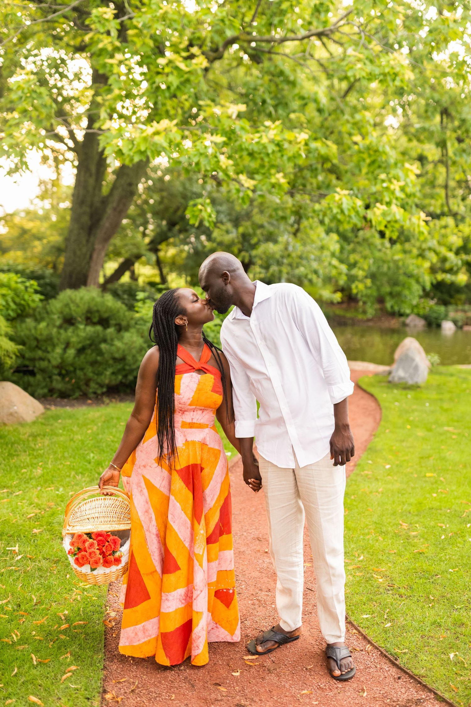 A couple shares a romantic kiss on a garden path, the woman holding a basket of flowers in vibrant attire.